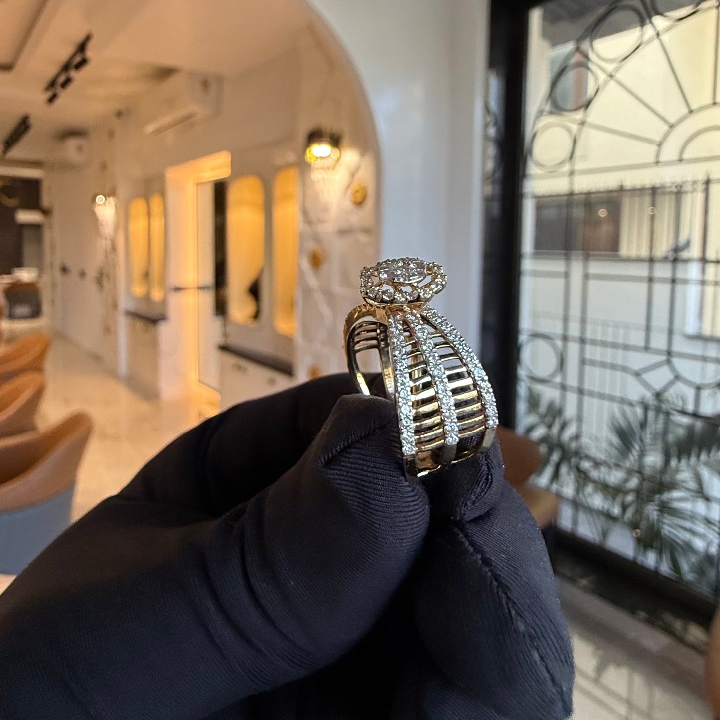 Elegant gold diamond ring featuring a round-cut central diamond surrounded by a halo of smaller diamonds, set on a split shank band adorned with rows of sparkling pavé diamonds, displayed on a black gloved hand against a neutral background.