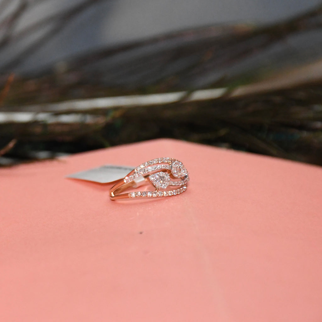 A rose gold ring with pavé-set clear diamonds, featuring a stylized curved design, resting on a pink surface with a blurred dark background.