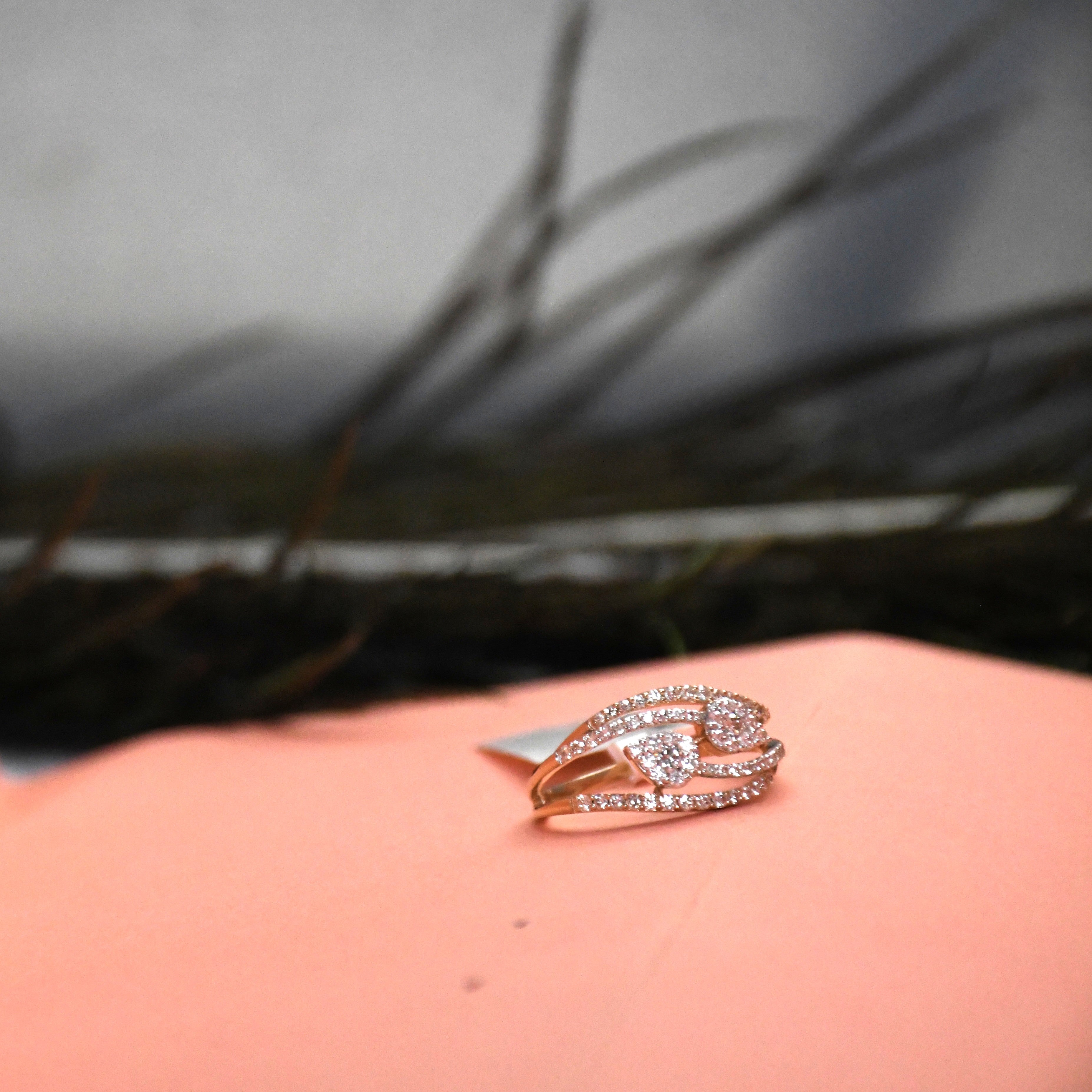 A rose gold ring with pavé-set clear diamonds, featuring a central pear-cut stone, resting on a peach-colored surface with a blurred dark background.