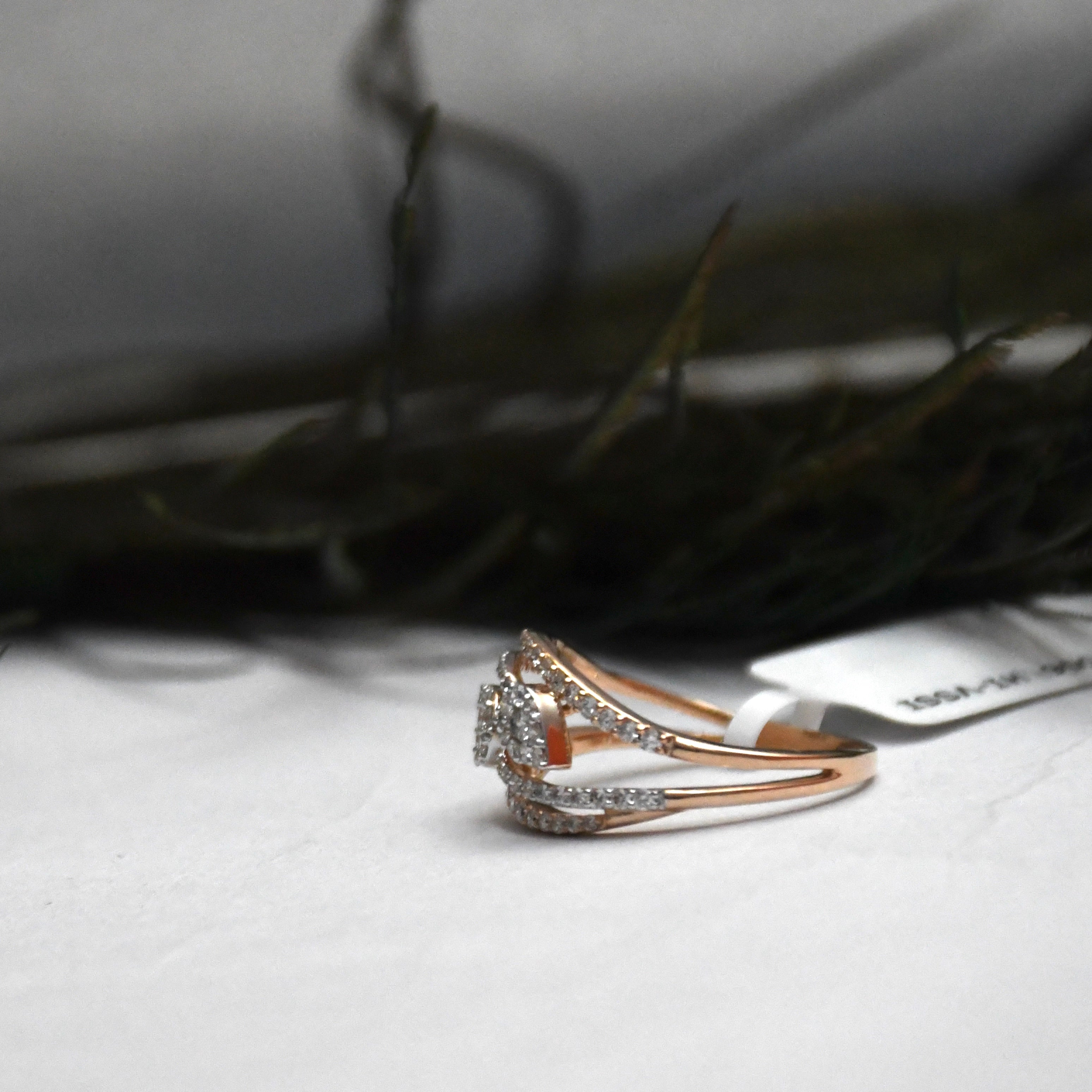 A rose gold ring with diamond cluster and pavé band, resting on a white surface with a blurred dark background.