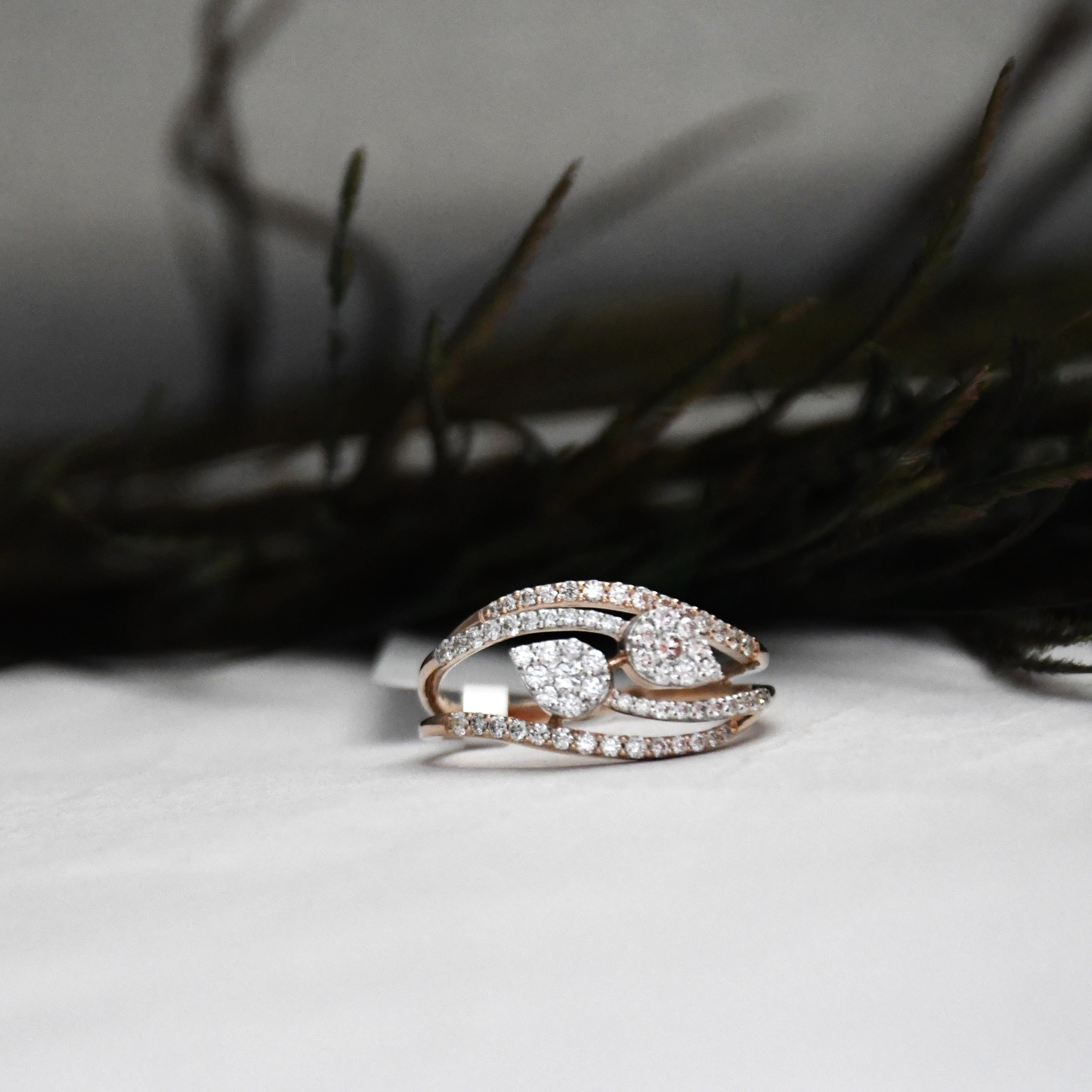 A rose gold ring with a pear-shaped diamond cluster center and pavé-set curved bands, resting on a white surface with dark, blurred foliage in the background.