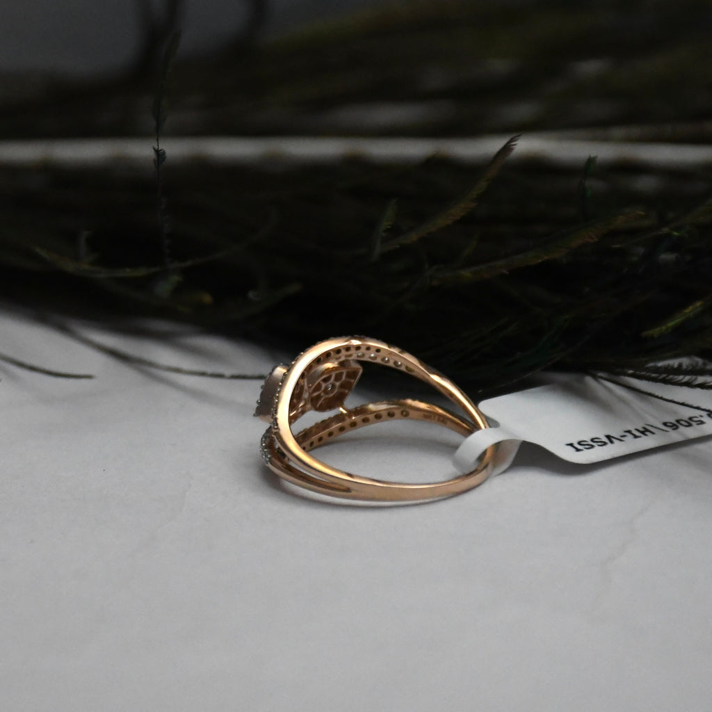A rose gold ring with a floral filigree setting and small clear diamonds, resting on a light gray surface with a white tag attached, against a dark, blurred background.