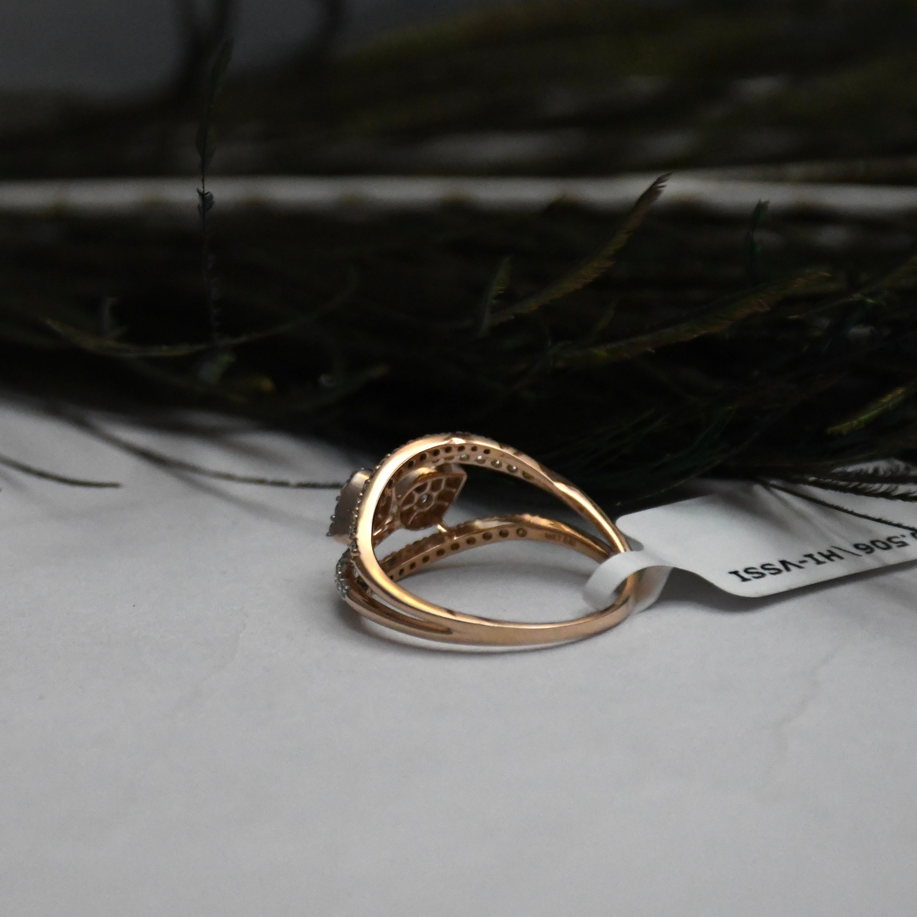 A rose gold ring with a floral filigree setting and small clear diamonds, resting on a light gray surface with a white tag attached, against a dark, blurred background.