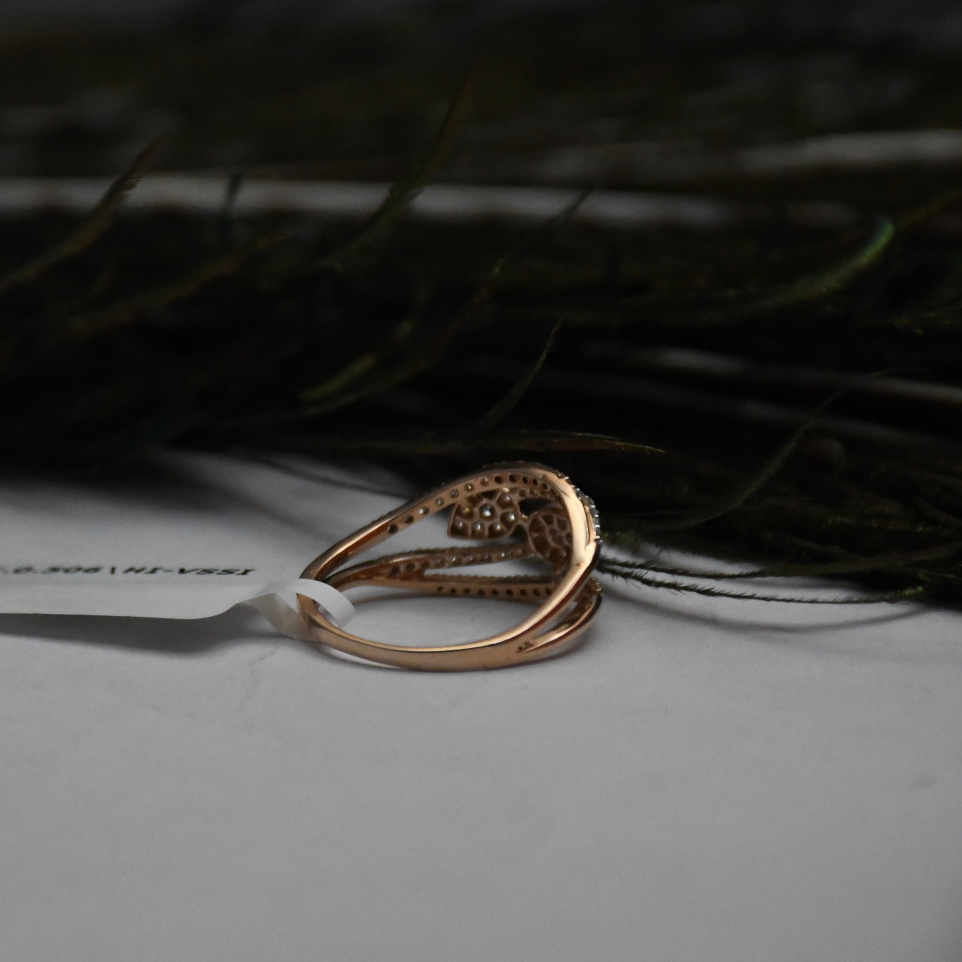 A rose gold ring with a textured, openwork design and a small central cluster of clear diamonds, resting on a white surface with a dark, blurred background.