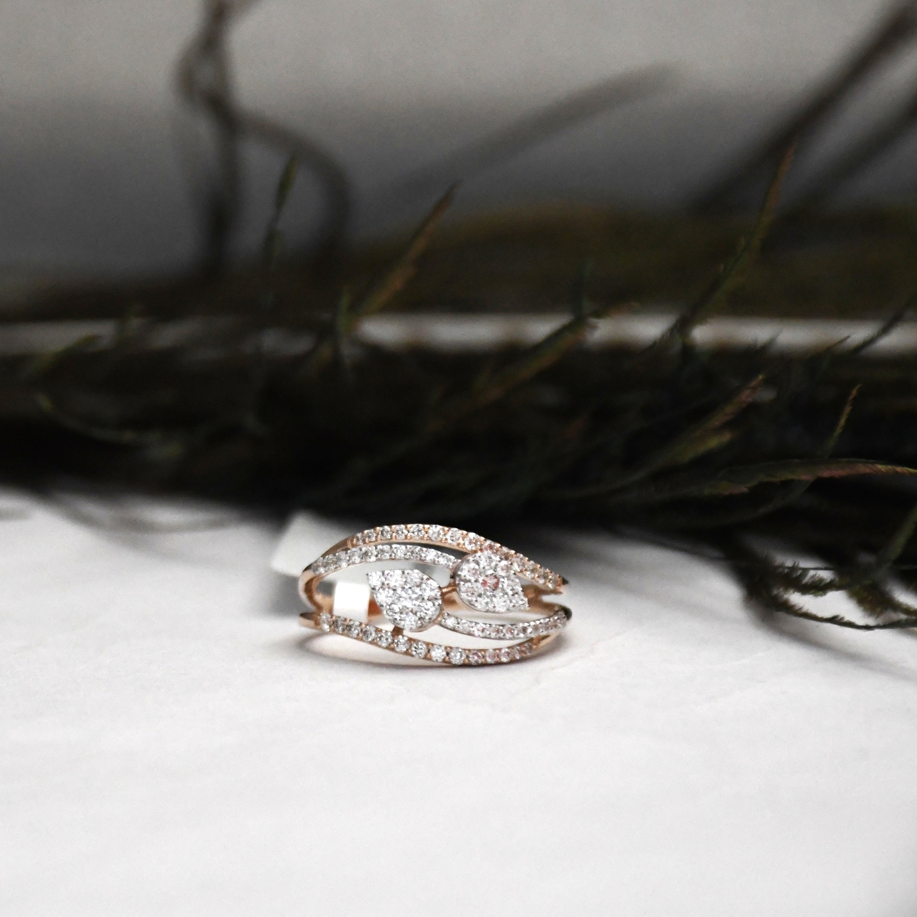 A rose gold ring with pavé-set clear stones, featuring a central pear shape cluster and four curved bands, resting on a white surface with dark blurred foliage in the background.
