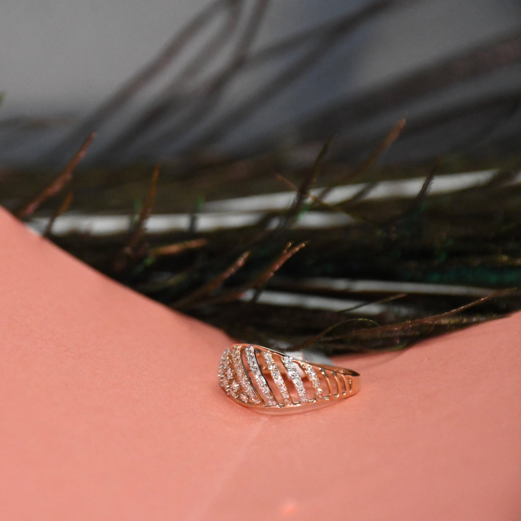 A rose gold ring with a leaf-like segmented design set with small clear stones, resting on a pink surface with dark feathers in the background.