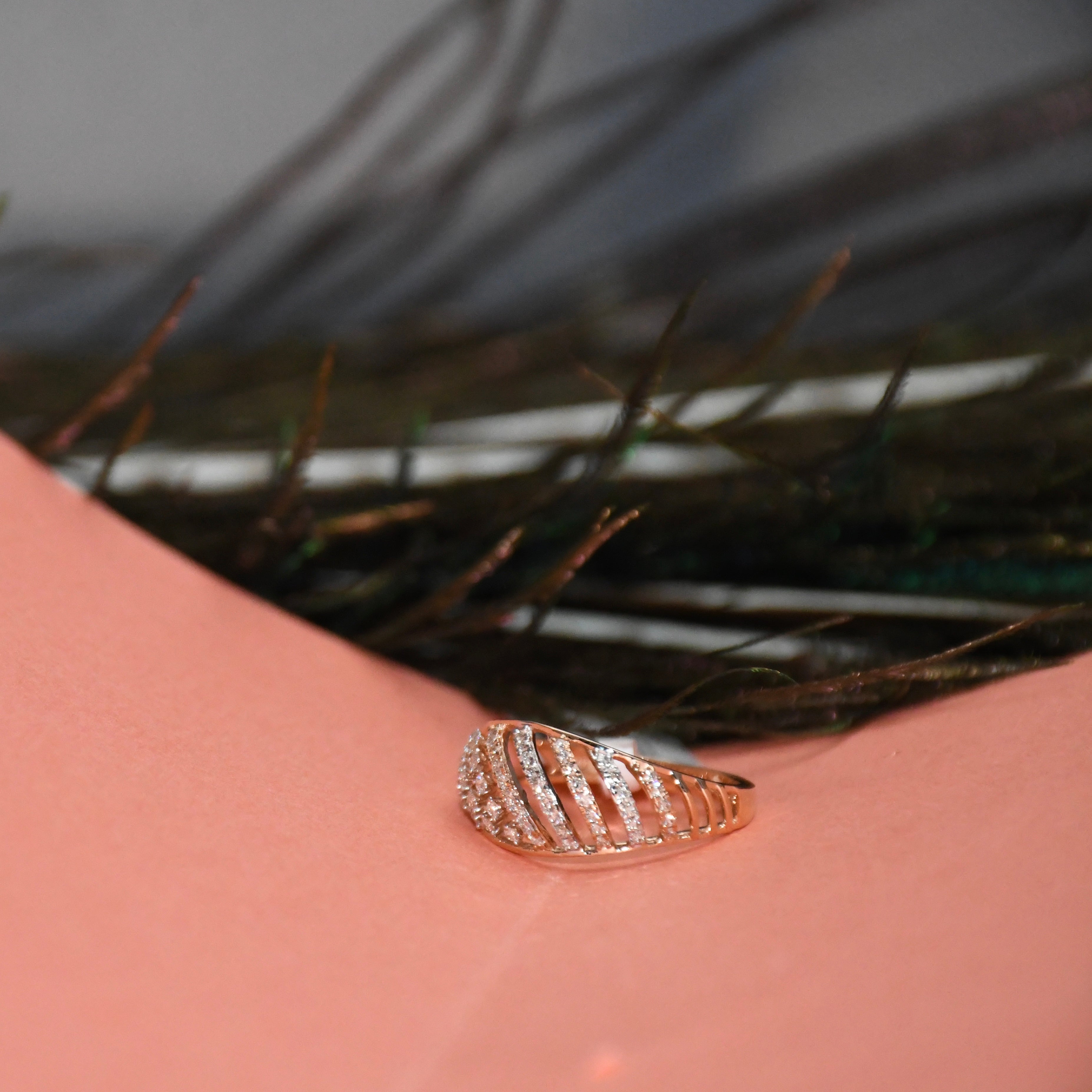 A rose gold ring with a leaf-like segmented design set with small clear stones, resting on a pink surface with dark feathers in the background.