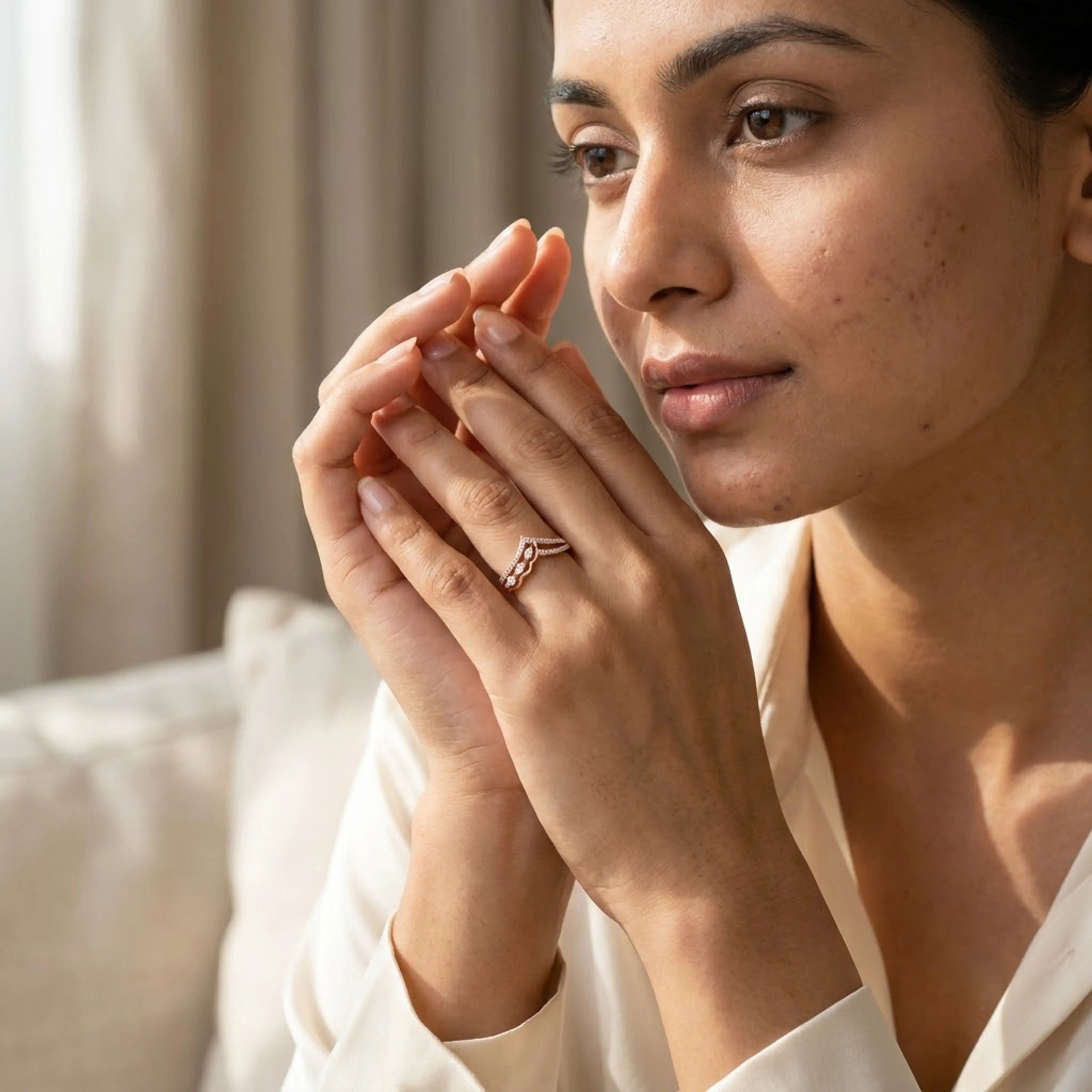 A close-up lifestyle shot of a woman wearing a delicate gold chevron ring on her finger, dressed in a white satin shirt against a soft, warm-toned indoor background.