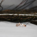 A rose gold ring with pavé-set clear diamonds, photographed on a white surface with dark dried grasses in the background.