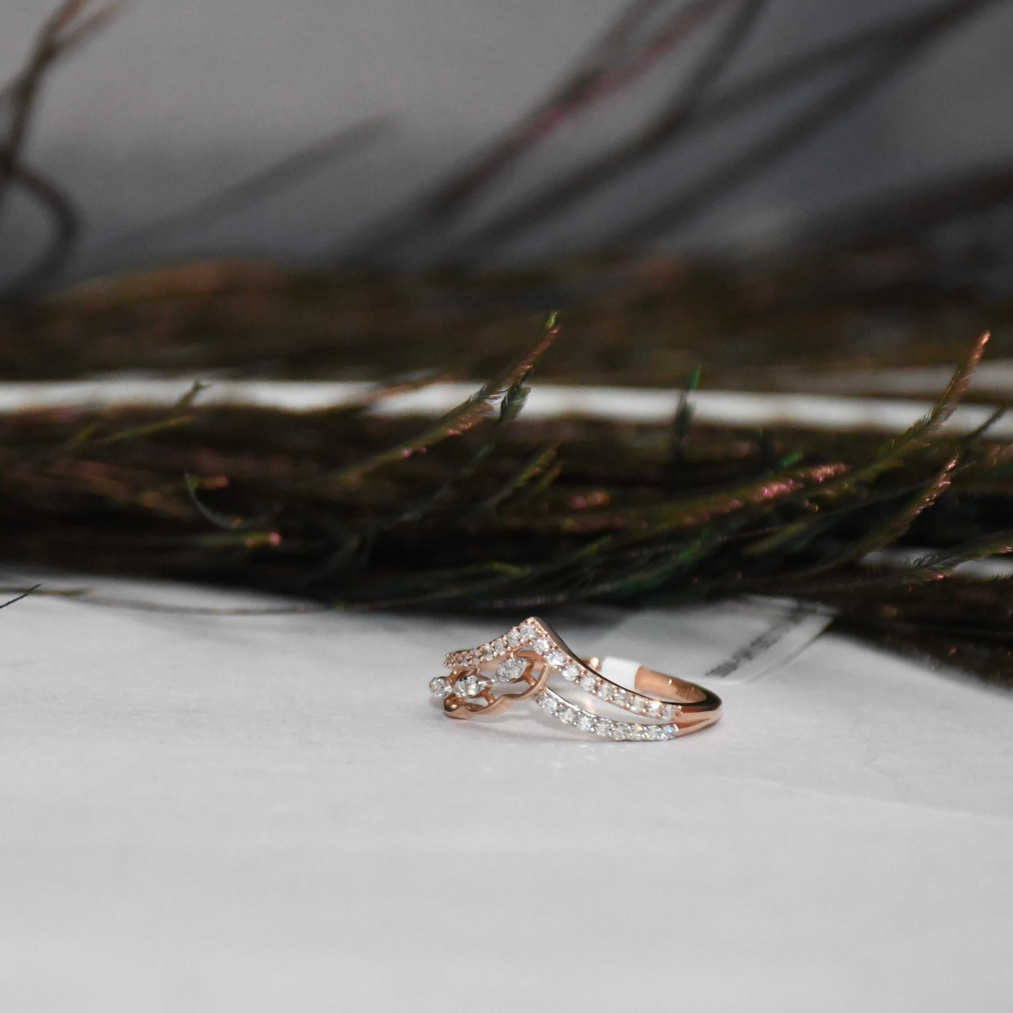 A rose gold ring with pavé-set clear diamonds, photographed on a white surface with dark dried grasses in the background.
