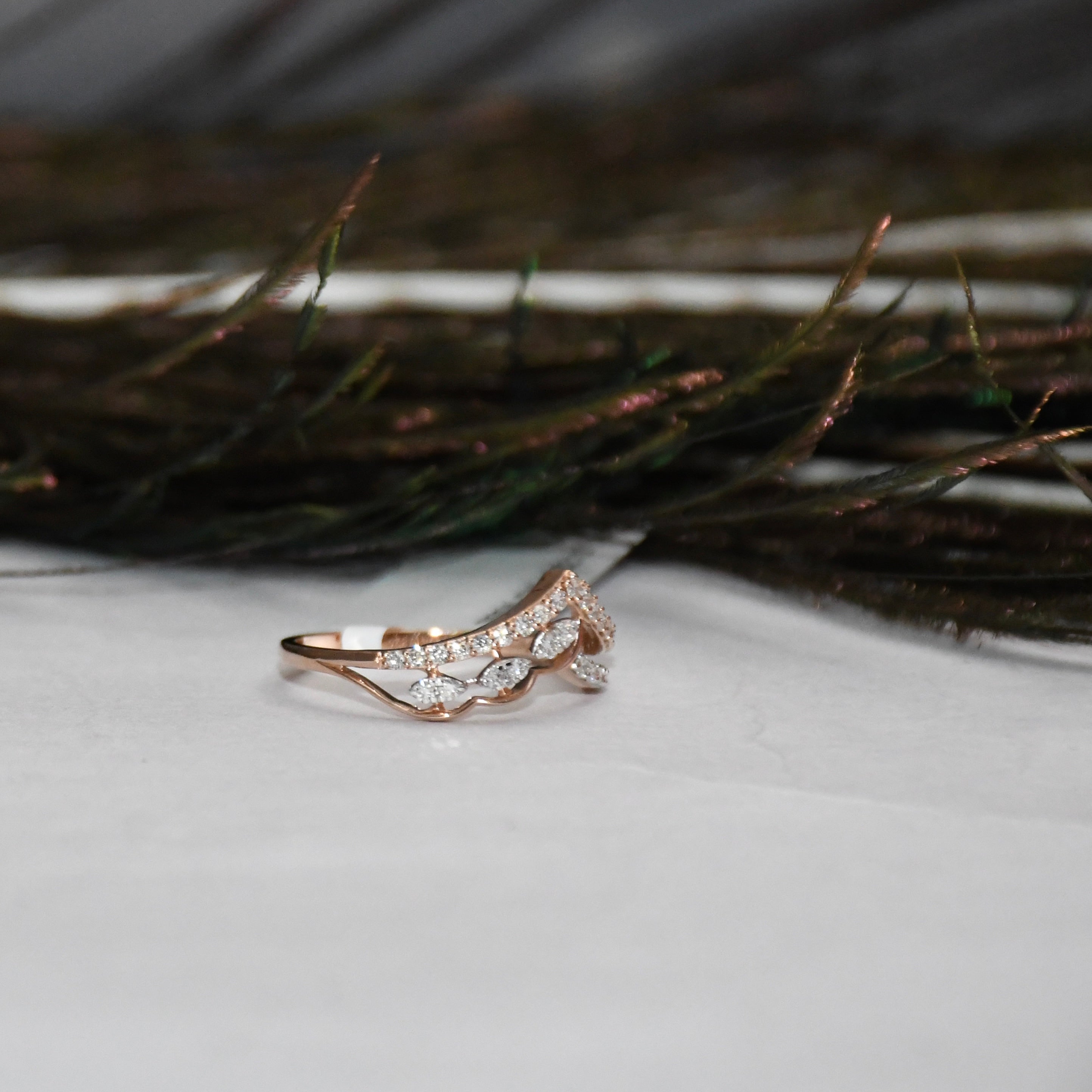 A rose gold ring with pavé-set clear stones, photographed on a white surface with dark feathers in the blurred background.