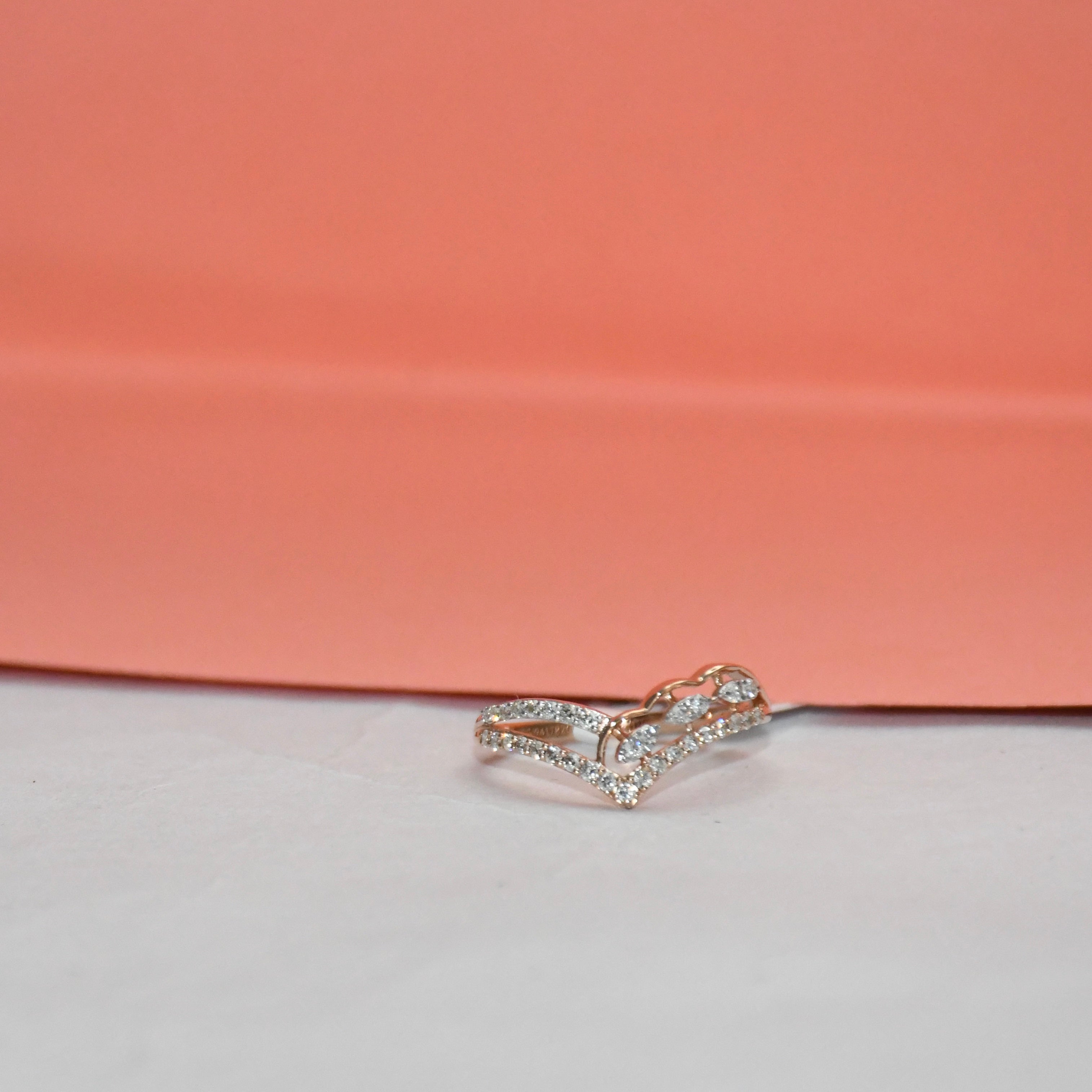 A delicate rose gold ring with pavé-set clear stones, photographed on a white surface with a peach-pink background.