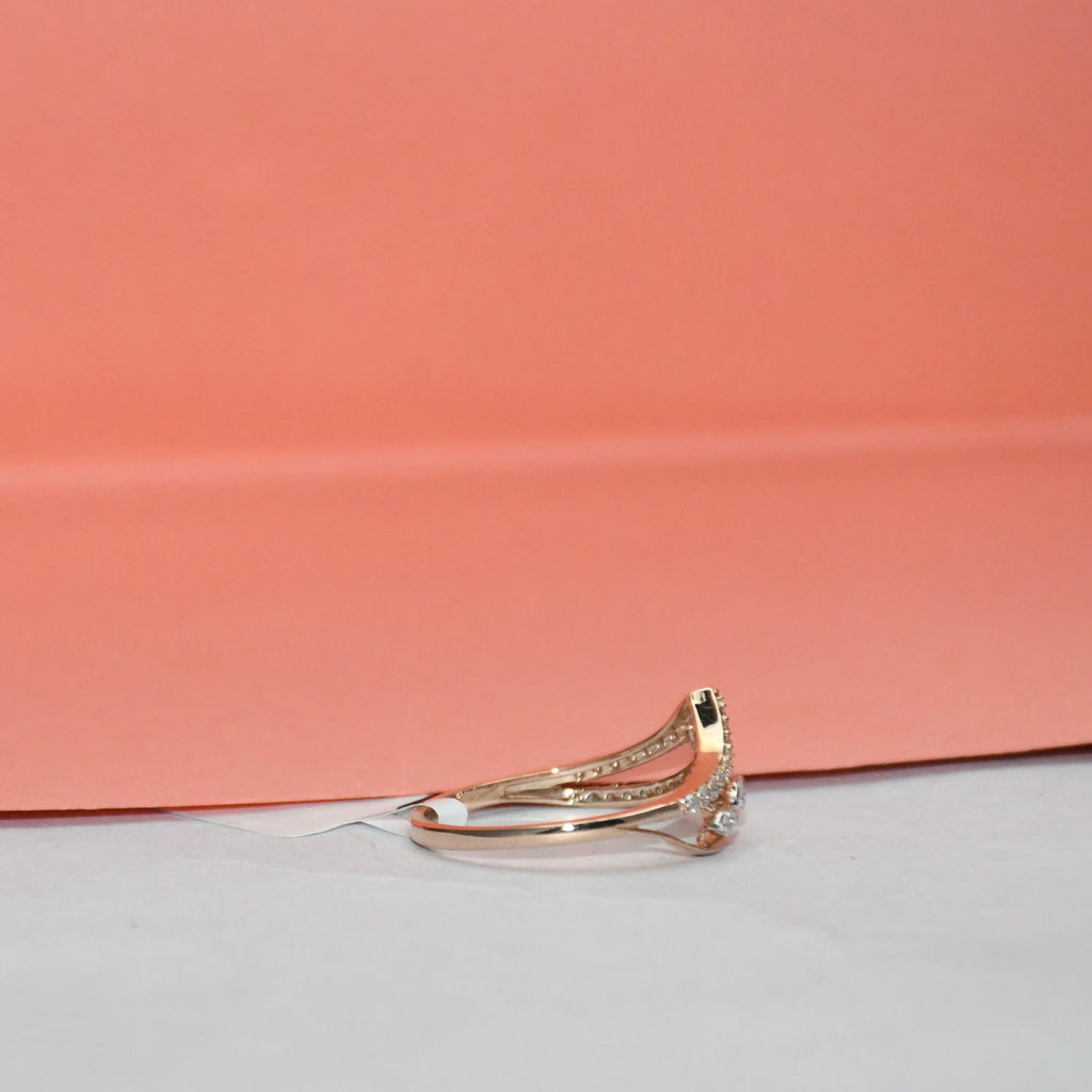 A gold ring with a curved pavé band and a small clear gemstone cluster on the front, photographed on a white surface with a peach-pink background.