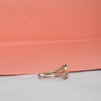 A gold ring with a curved pavé band and a small clear gemstone cluster on the front, photographed on a white surface with a peach-pink background.