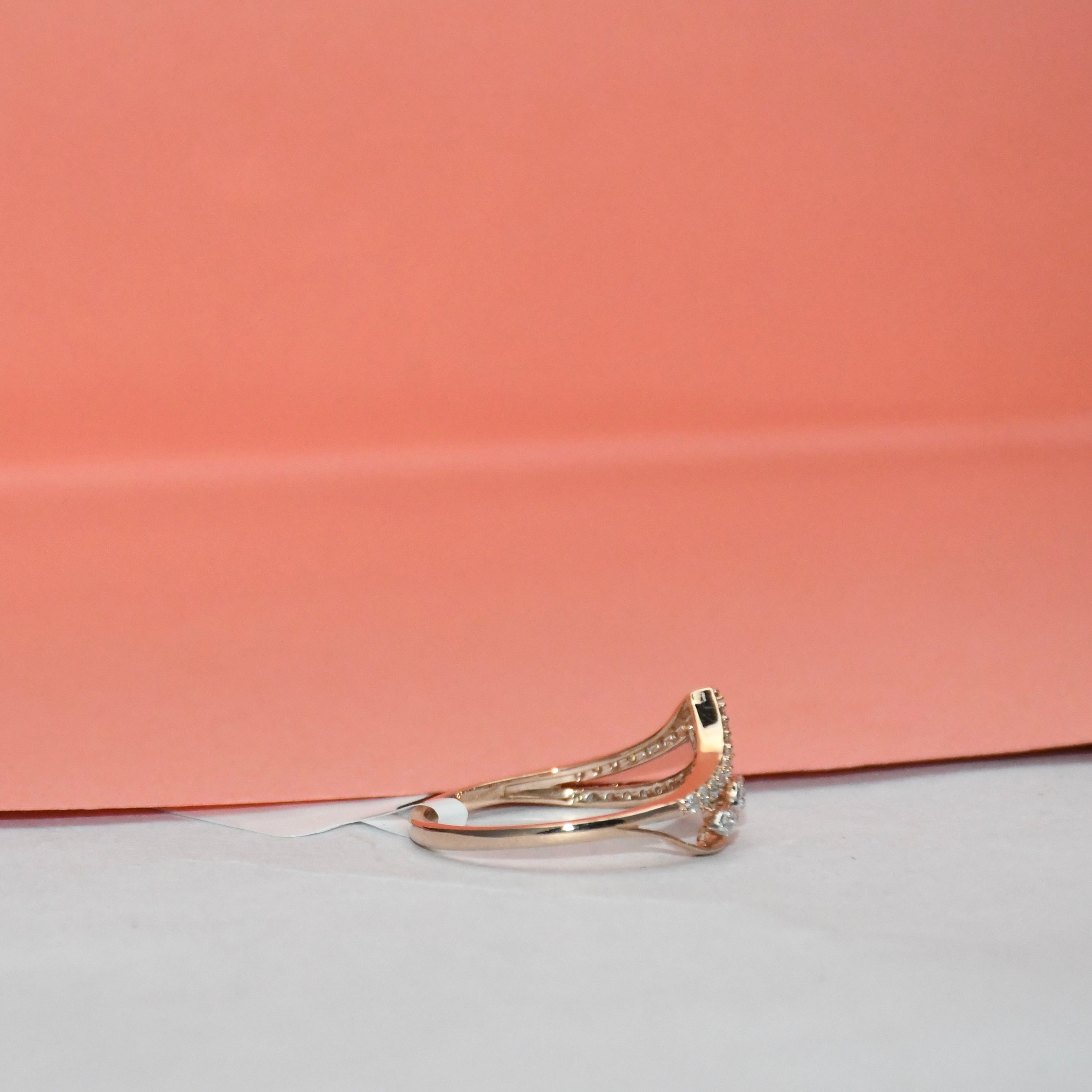A gold ring with a curved pavé band and a small clear gemstone cluster on the front, photographed on a white surface with a peach-pink background.