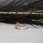A rose gold ring with an intricate filigree top, resting on a white surface with dark decorative grass in the background.
