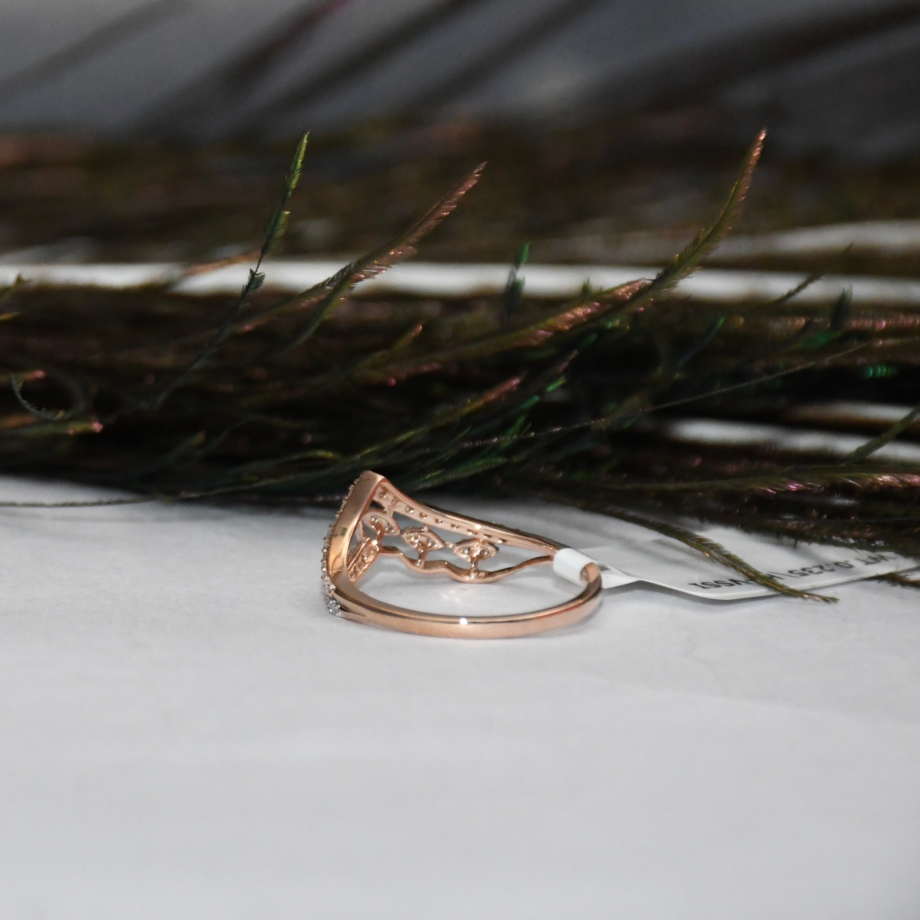 A rose gold ring with an intricate filigree top, resting on a white surface with dark decorative grass in the background.