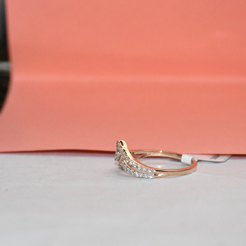 A delicate curved ring featuring a pavé diamond setting in gold metal and a rose gold accent on the band, resting on a light gray surface with a peach-colored background.
