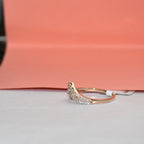 A delicate curved ring featuring a pavé diamond setting in gold metal and a rose gold accent on the band, resting on a light gray surface with a peach-colored background.