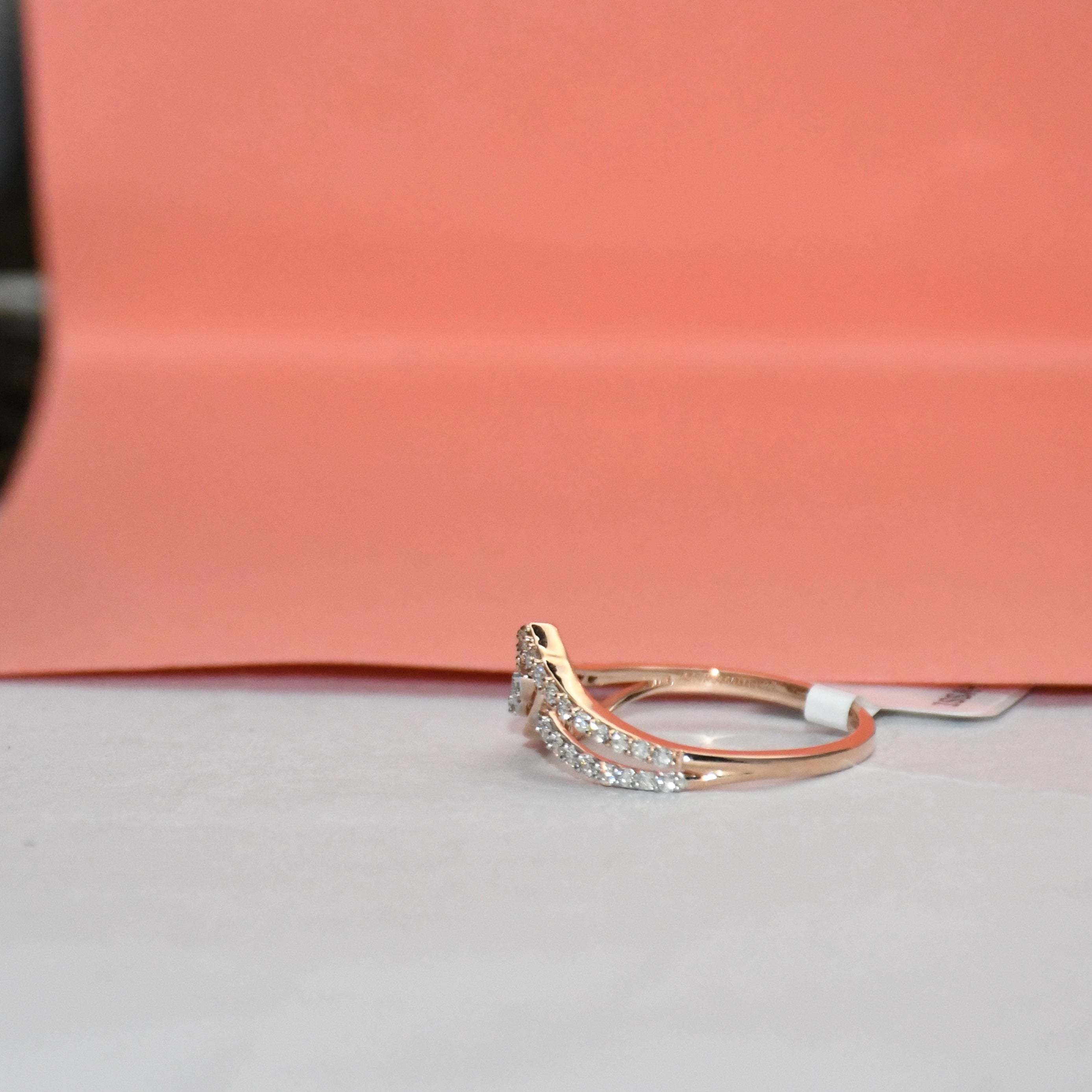 A delicate curved ring featuring a pavé diamond setting in gold metal and a rose gold accent on the band, resting on a light gray surface with a peach-colored background.