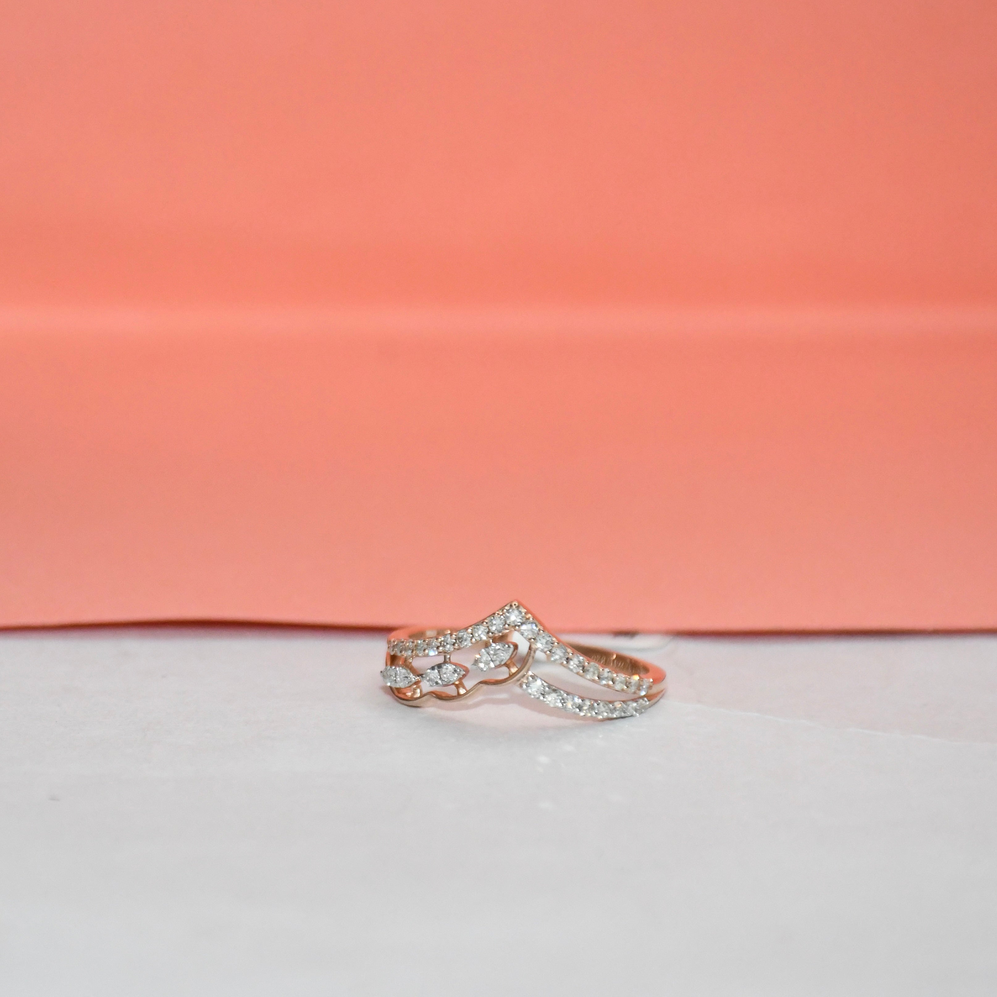 A rose gold ring with pavé-set clear stones, photographed on a white surface with a peach-pink background.