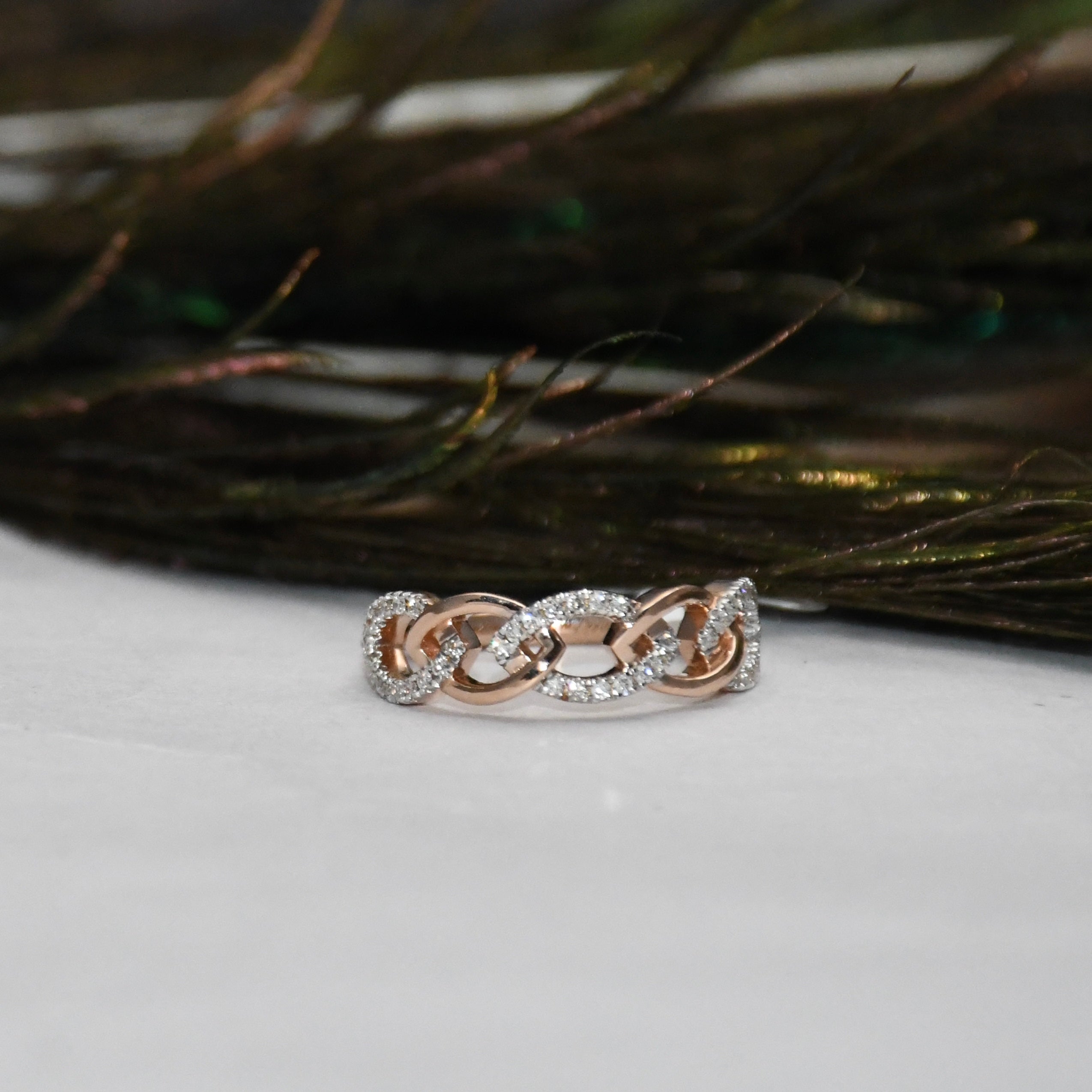 A delicate chain-link ring in rose gold, accented with small clear pavé stones, resting on a white surface with dark feathers in the background.