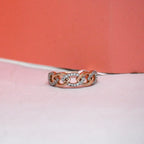 A rose gold chain-link ring with pavé-set clear stones, resting on a light pink surface with a coral-pink background.