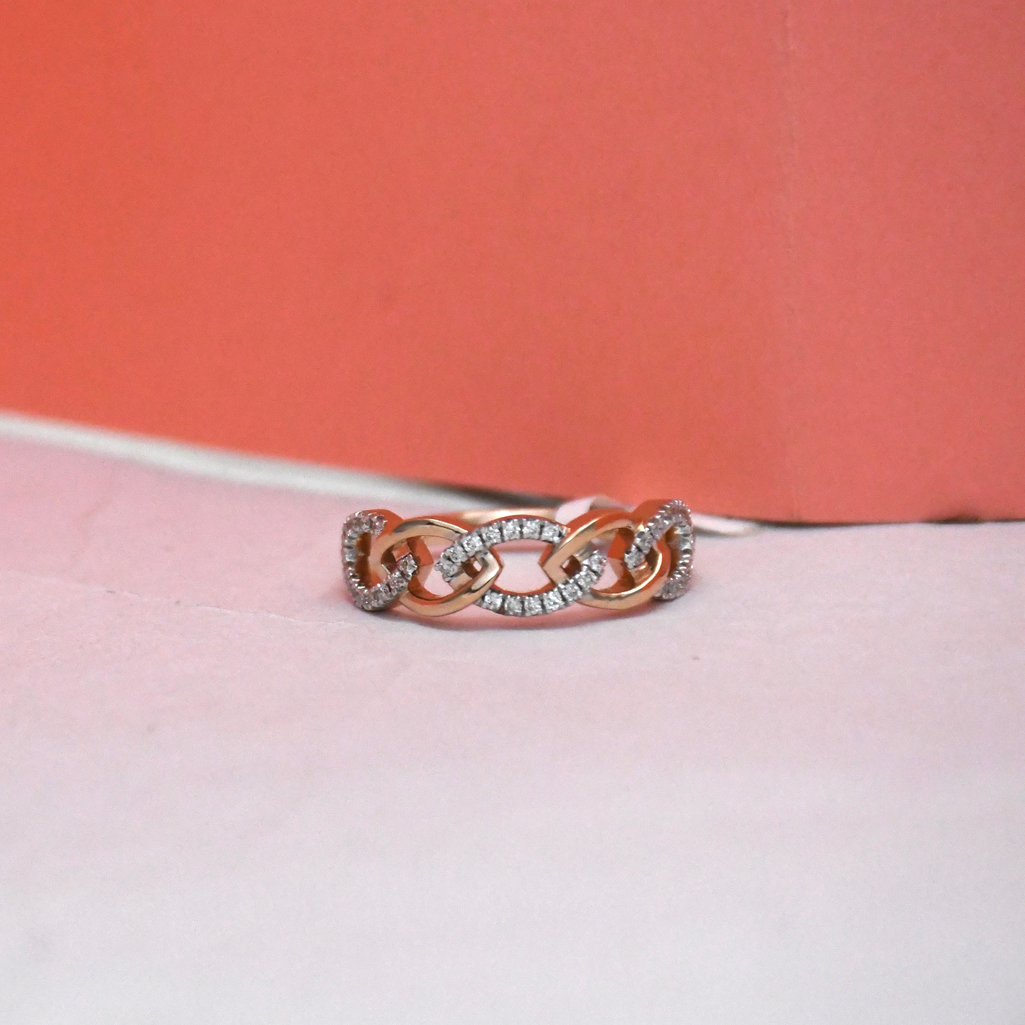 A rose gold chain-link ring with pavé-set clear stones, resting on a light pink surface with a coral-pink background.