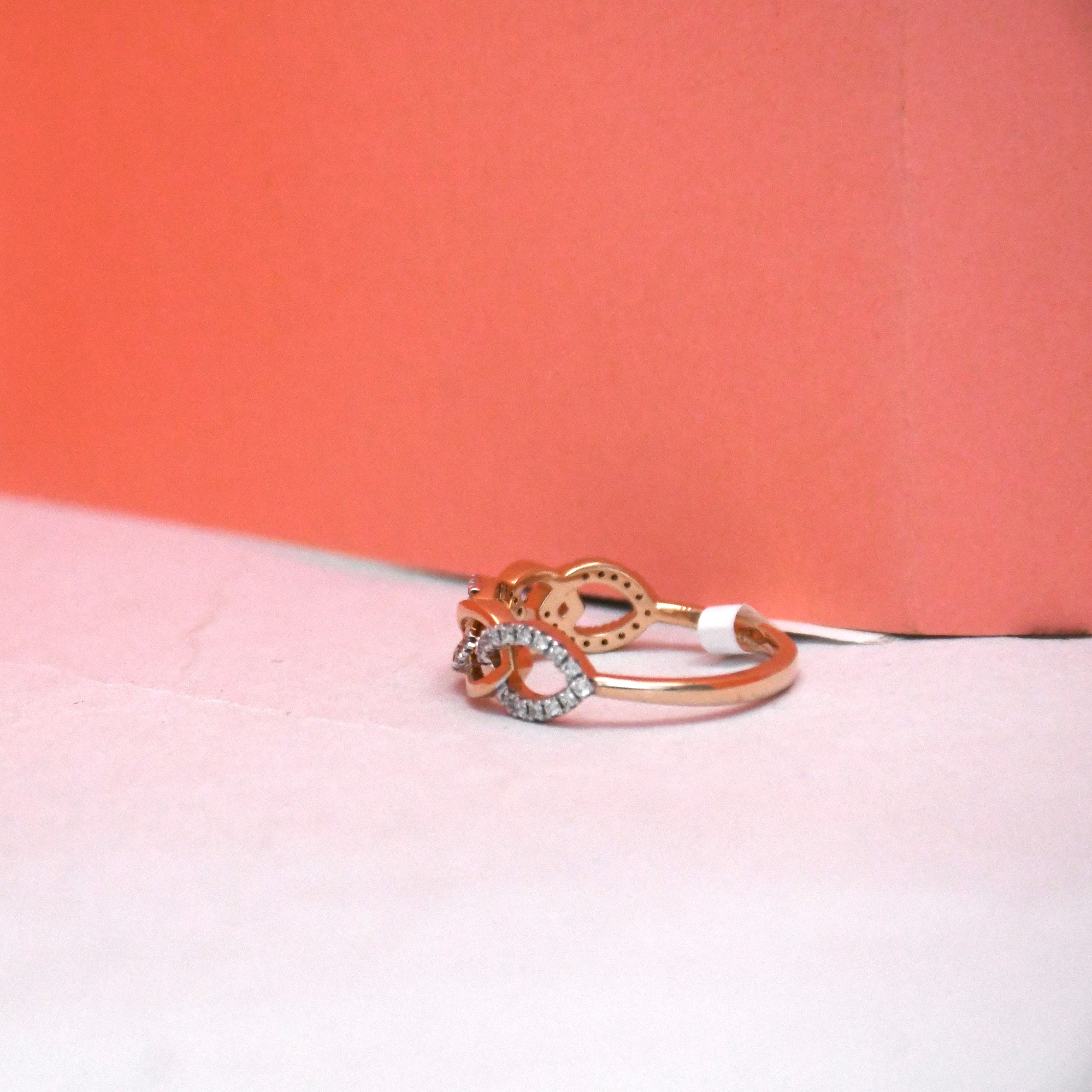 A rose gold ring with a pavé setting, resting on a light pink surface with a coral-pink background box.