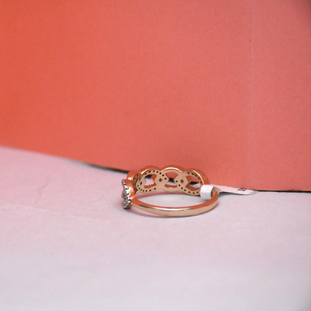 A rose gold ring with a decorative openwork top and a small clear gemstone on the band, resting on a light surface against a coral background.
