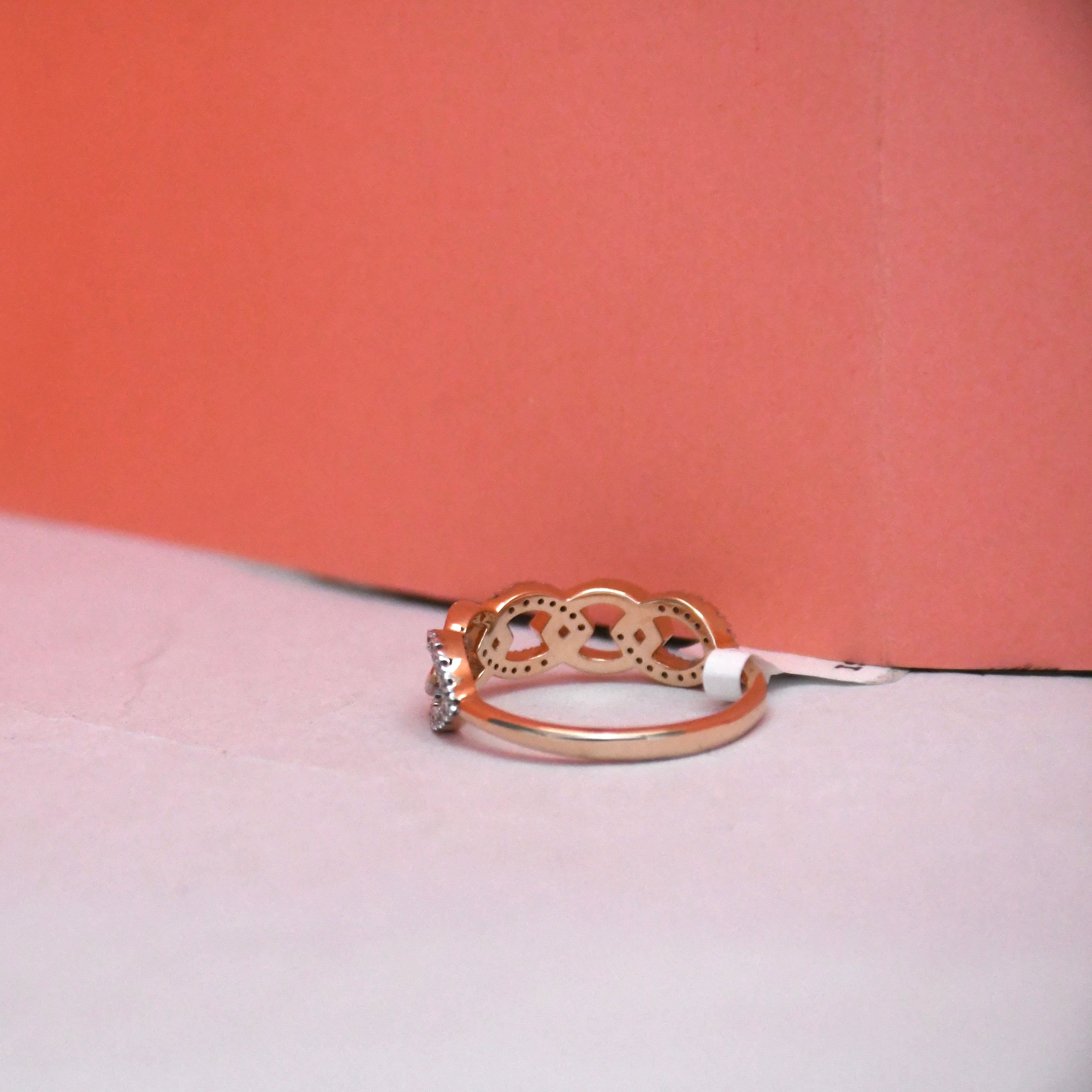 A rose gold ring with a decorative openwork top and a small clear gemstone on the band, resting on a light surface against a coral background.