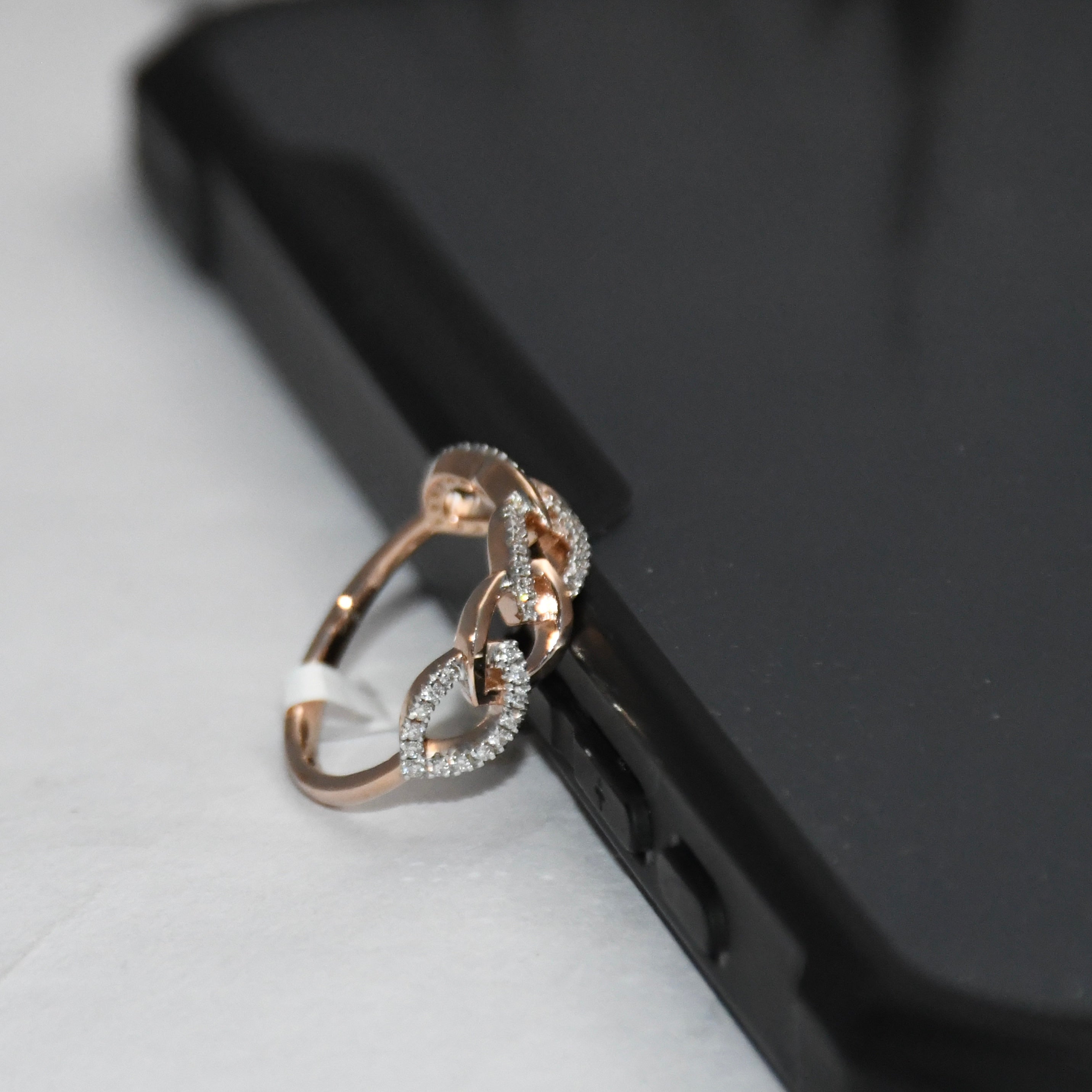 A rose gold ring with interlocking chain-link designs and pavé-set clear stones, resting on the edge of a black smartphone against a light gray background.