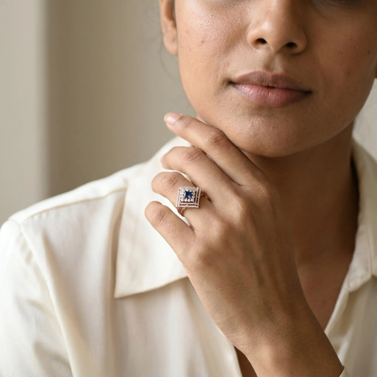 A close-up lifestyle shot of a model wearing a square ring with a deep blue center stone and a halo of small clear stones in a rose gold setting, paired with a cream-colored collared shirt.