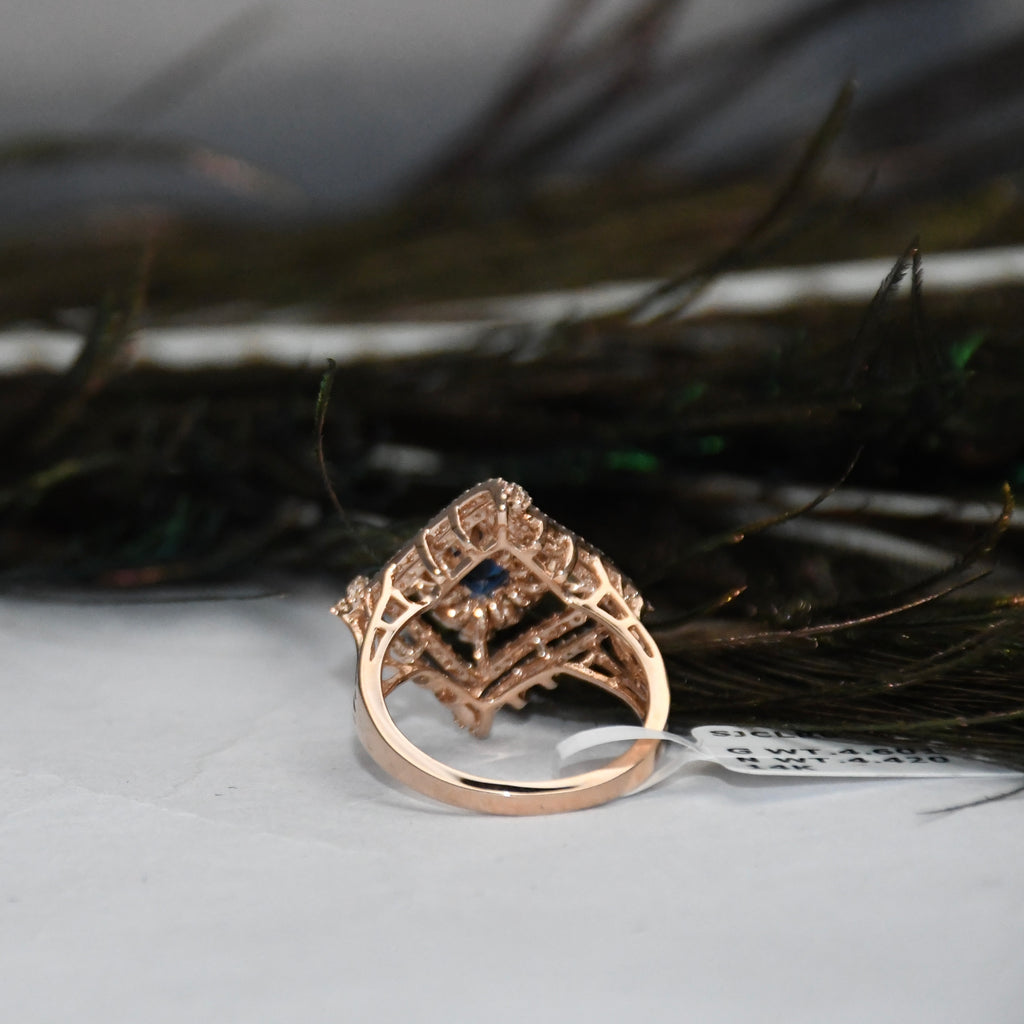 Backside of a rose gold ring with a geometric, basketweave-like openwork design and a small dark blue stone in the center, resting on a white surface with dark feathers in the background.
