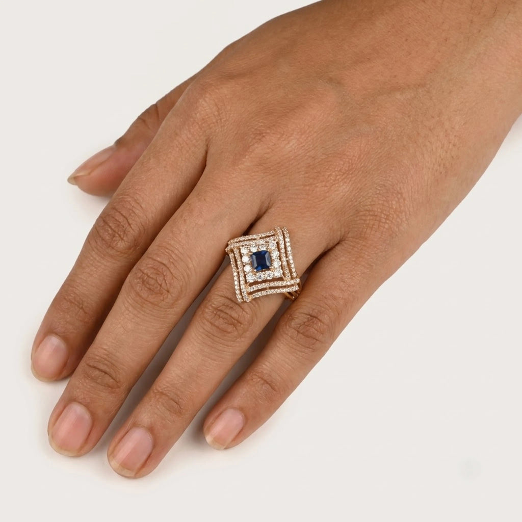 A close-up of a hand wearing a square ring with a central blue gemstone and surrounding small clear stones on a plain light background.