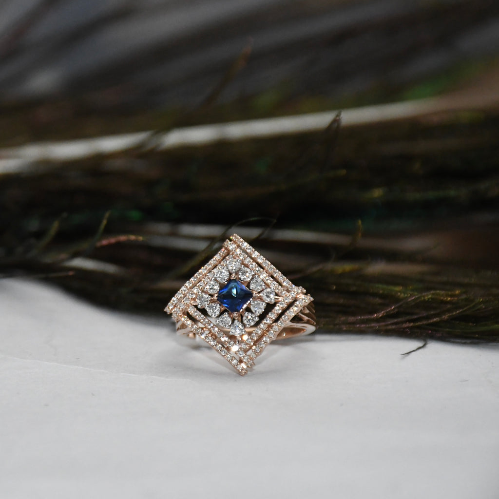 A square diamond-shaped ring with a central blue gemstone and surrounding pavé-set clear stones, displayed on a white surface with dark feather-like decor in the background.