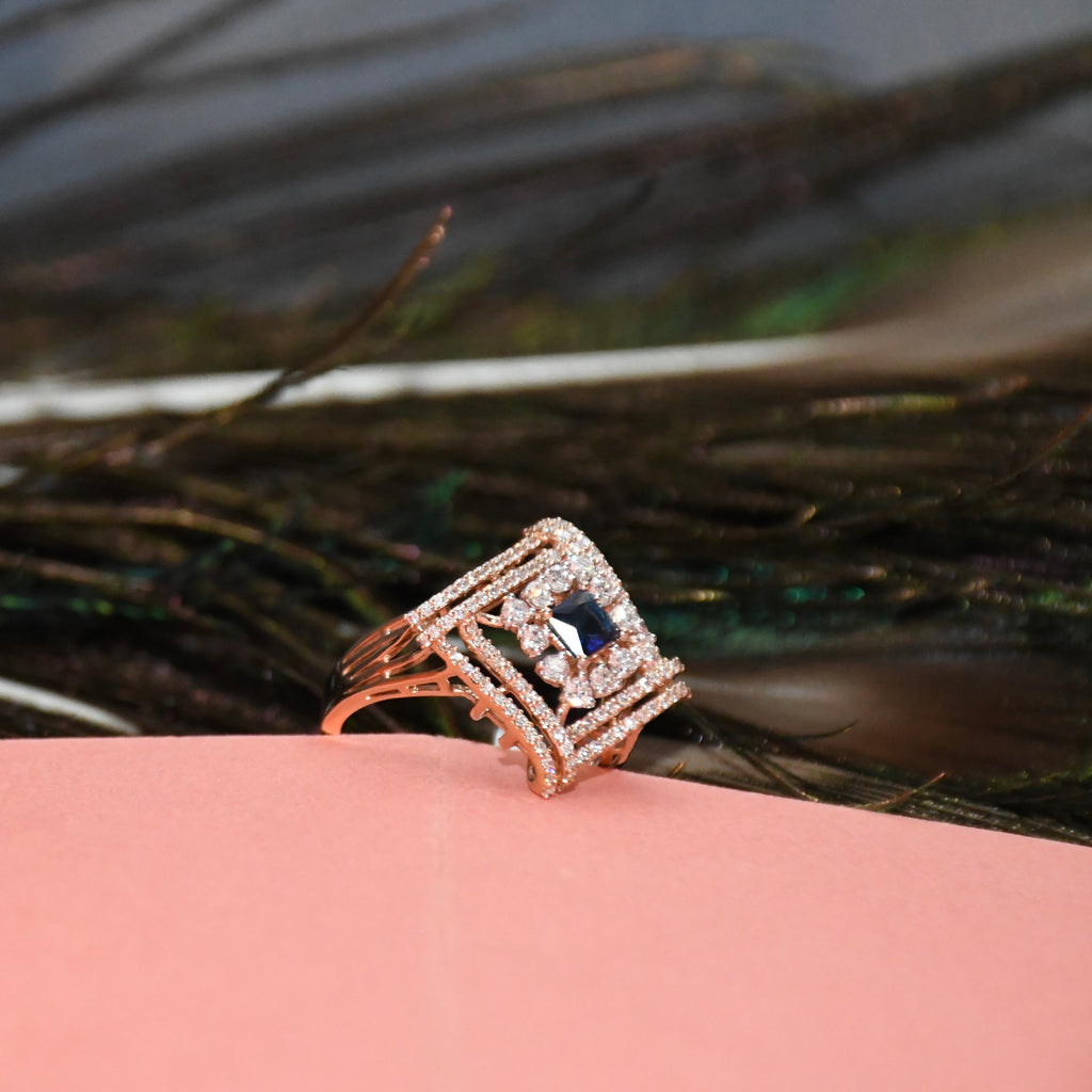 A rose gold ring with a square-cut dark blue center stone and surrounding pavé accents, resting on a pink surface with dark feathers in the background.