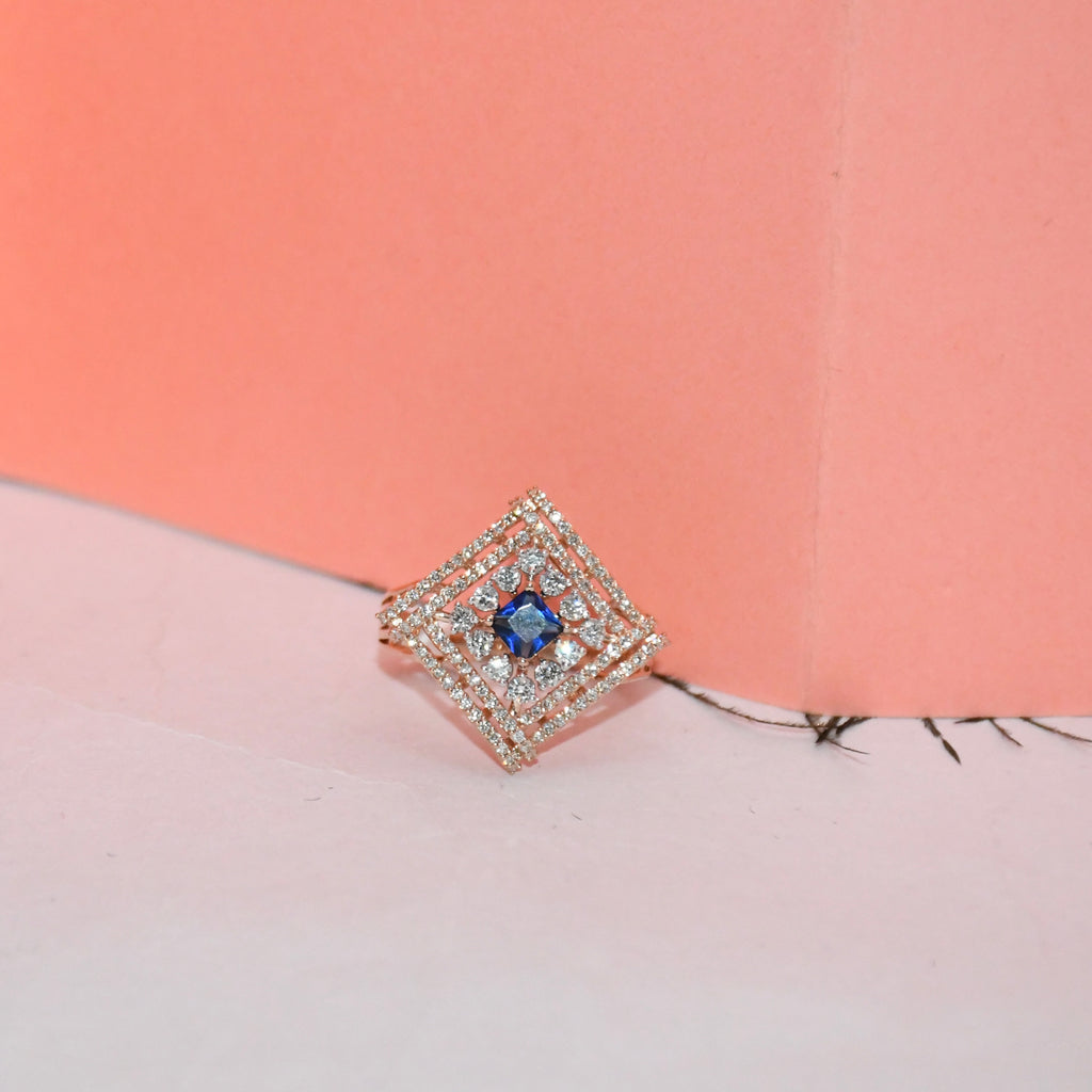 A square diamond-shaped ring featuring a central blue gemstone surrounded by a halo of small clear stones in a rose gold setting, displayed on a pink and peach background.