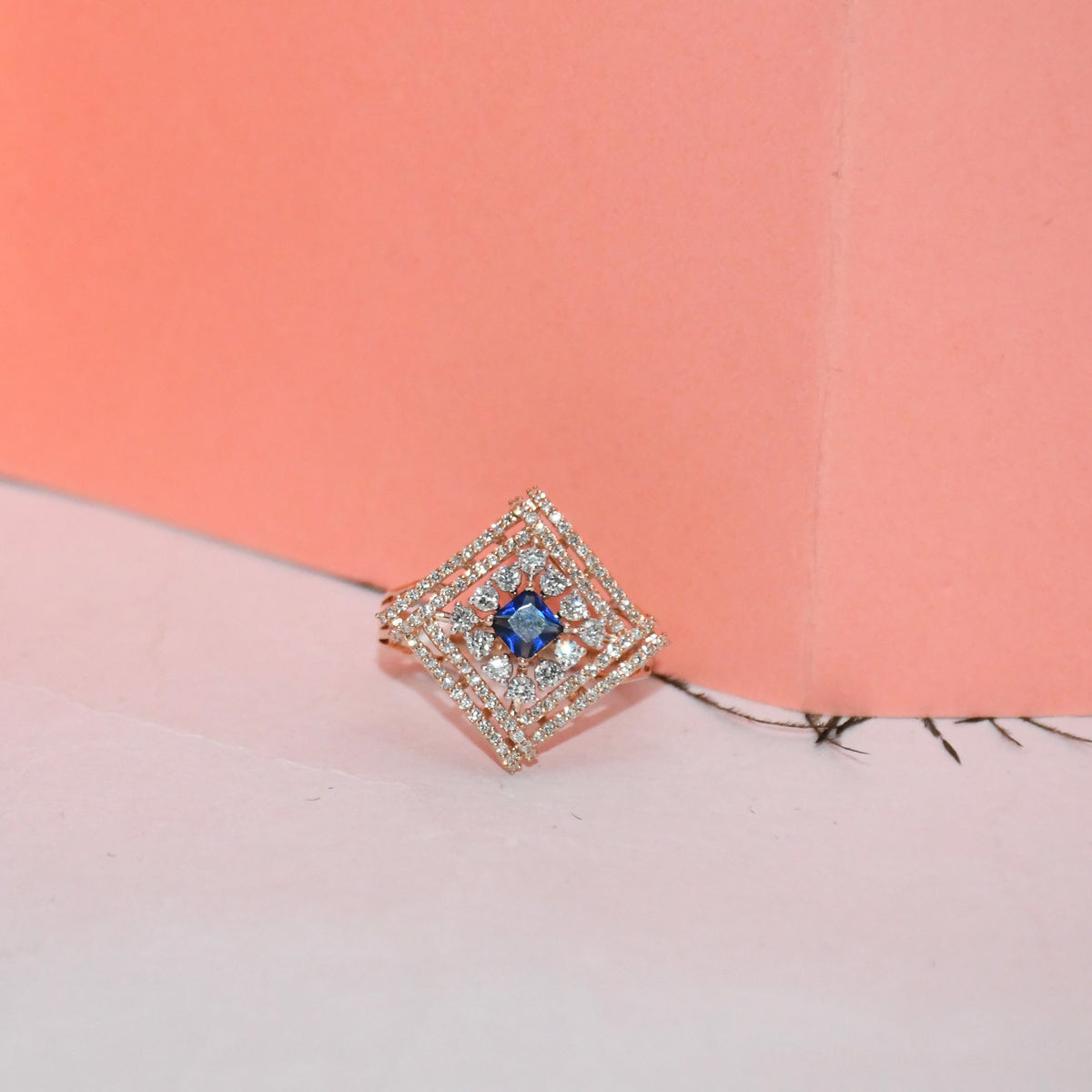 A square diamond-shaped ring featuring a central blue gemstone surrounded by a halo of small clear stones in a rose gold setting, displayed on a pink and peach background.
