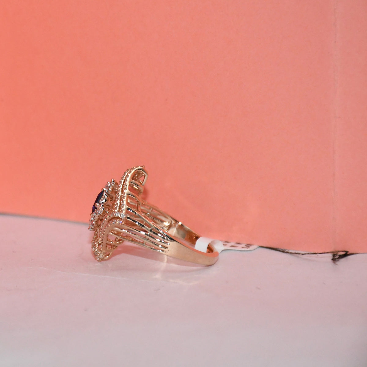 A gold ring with a decorative filigree band and a central dark blue stone, resting on a white surface against a peach-pink background.