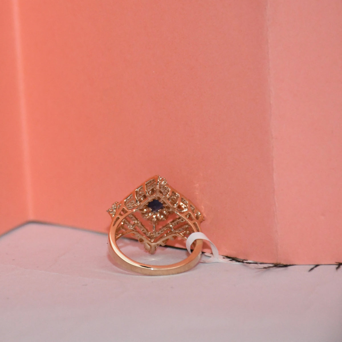 A rose gold ring with a decorative filigree top and a central dark blue stone, resting on a white surface against a coral pink background.