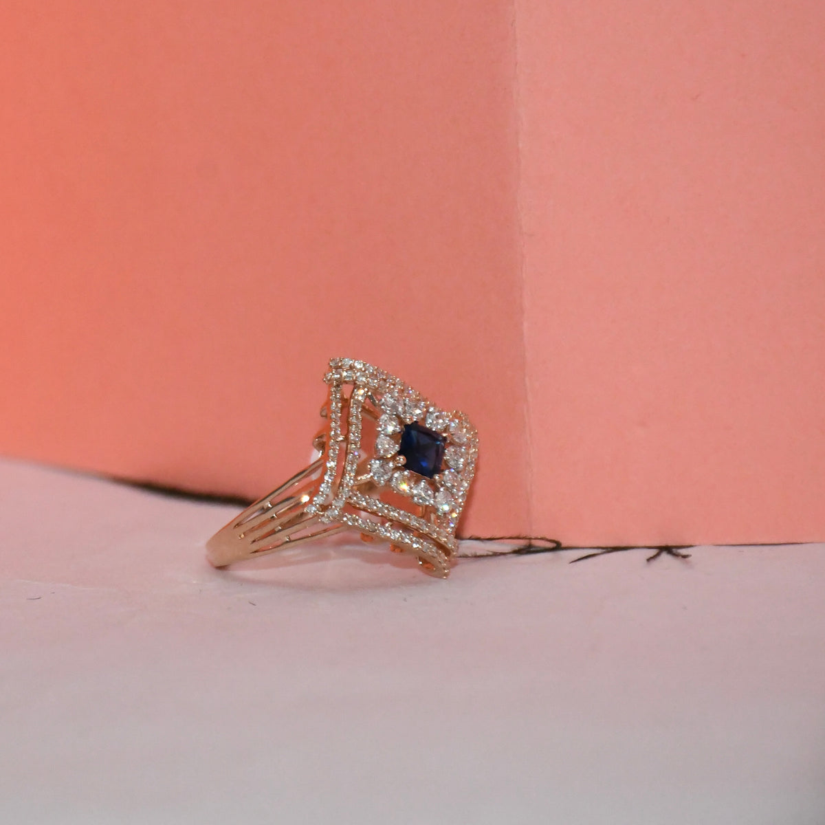 A rose gold ring with a square-shaped blue sapphire center stone and surrounding pavé diamonds, resting on a light pink surface against a coral pink background.