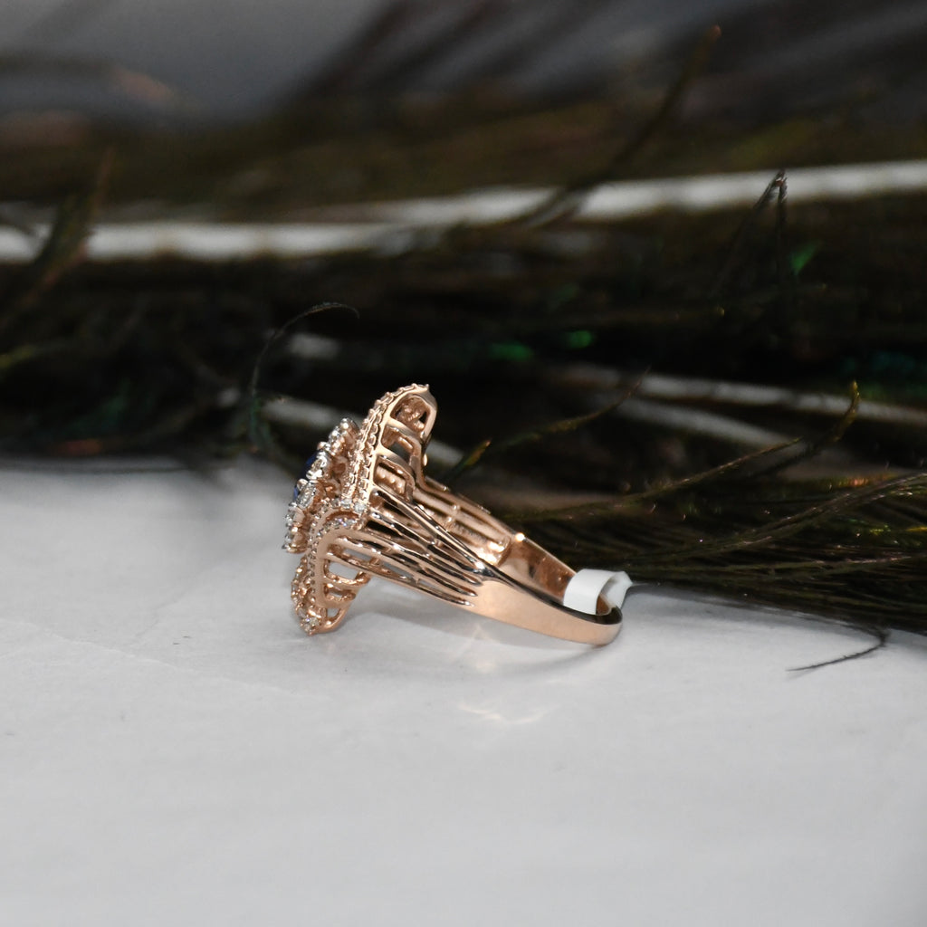 A rose gold ring with a floral cluster setting and small clear stones, resting on a white surface with dark feathers in the background.