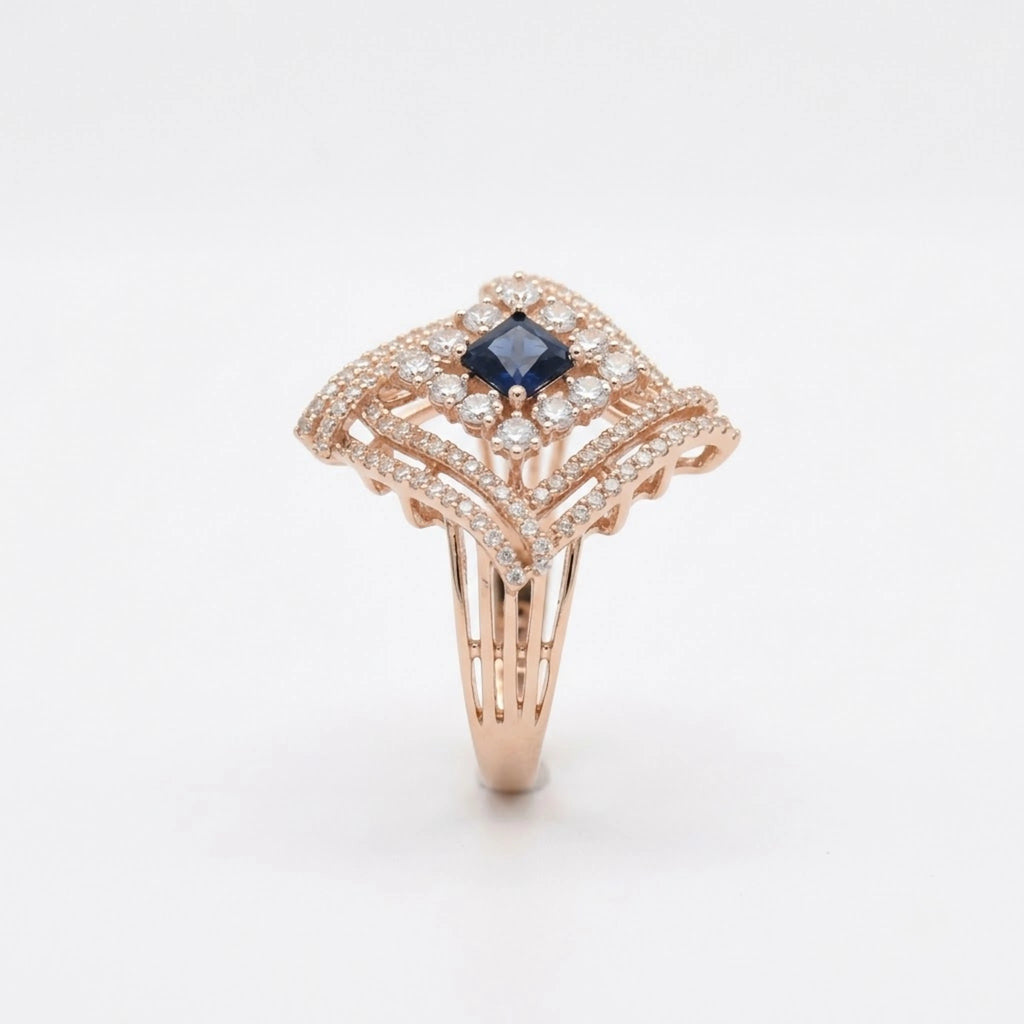 A rose gold ring featuring a central princess-cut dark blue sapphire surrounded by pavé-set clear diamonds, with an Art Deco-inspired openwork design on a clean white background.