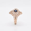 A rose gold ring featuring a central princess-cut dark blue sapphire surrounded by pavé-set clear diamonds, with an Art Deco-inspired openwork design on a clean white background.