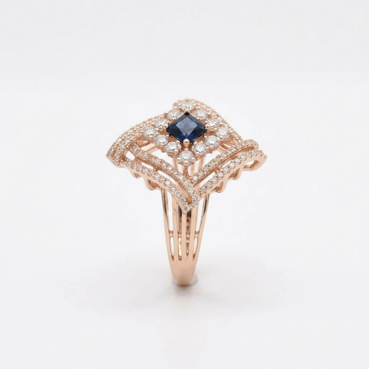 A rose gold ring featuring a central princess-cut dark blue sapphire surrounded by pavé-set clear diamonds, with an Art Deco-inspired openwork design on a clean white background.