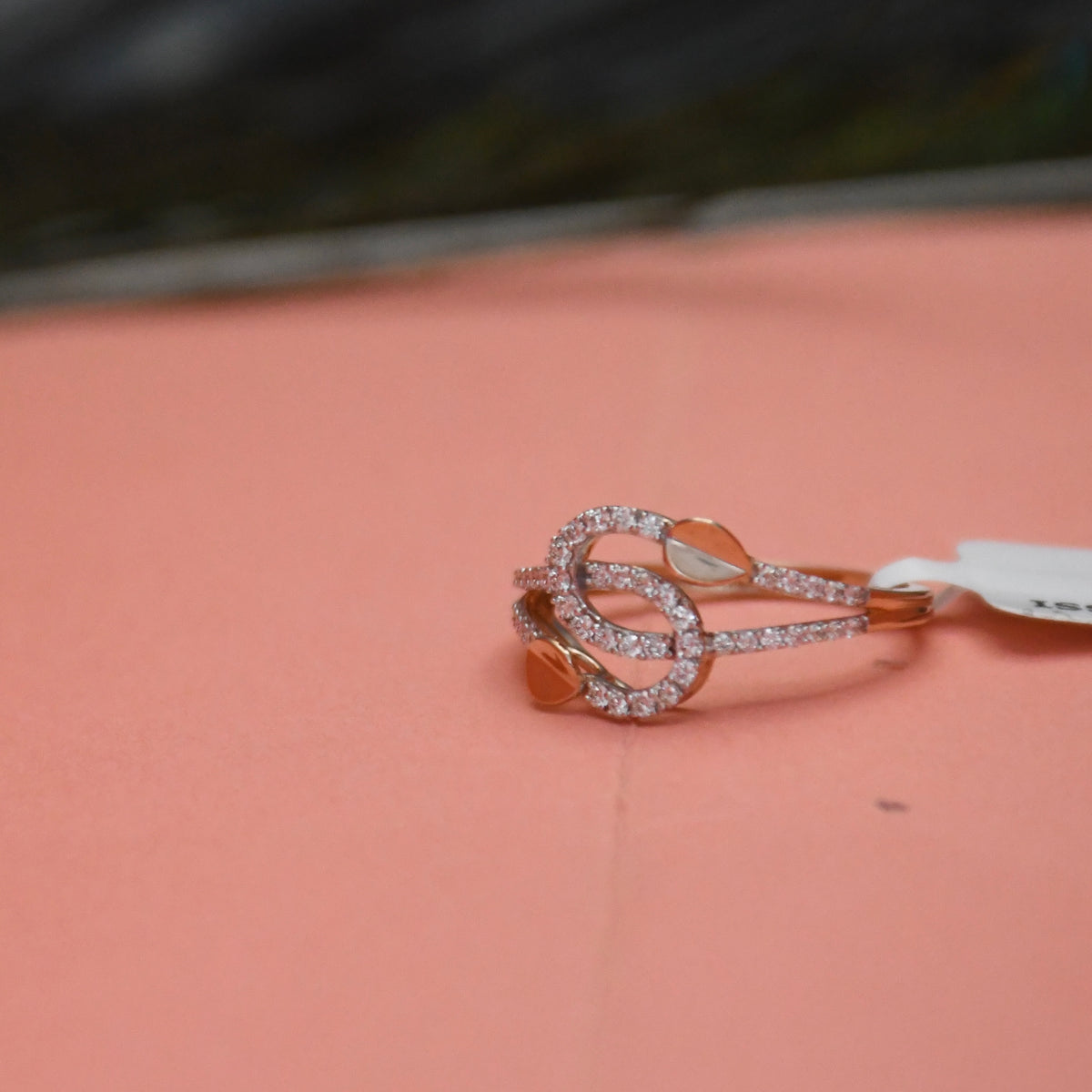 A rose gold ring with a double knot design, embellished with pavé diamonds, resting on a dusty pink surface.