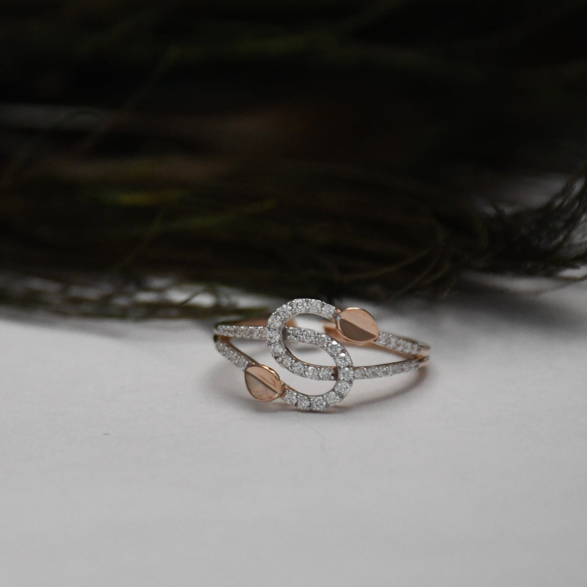 A delicate ring with intertwined gold bands, accented by small pavé-set clear stones and two gold shaped details, resting on a light gray surface with a dark, blurred background.