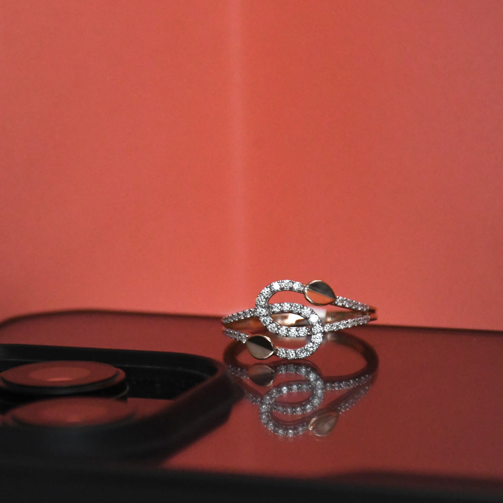 A gold toned ring adorned with pavé-set clear stones and smooth metallic scales, resting on a reflective dark surface against a warm coral-pink background.