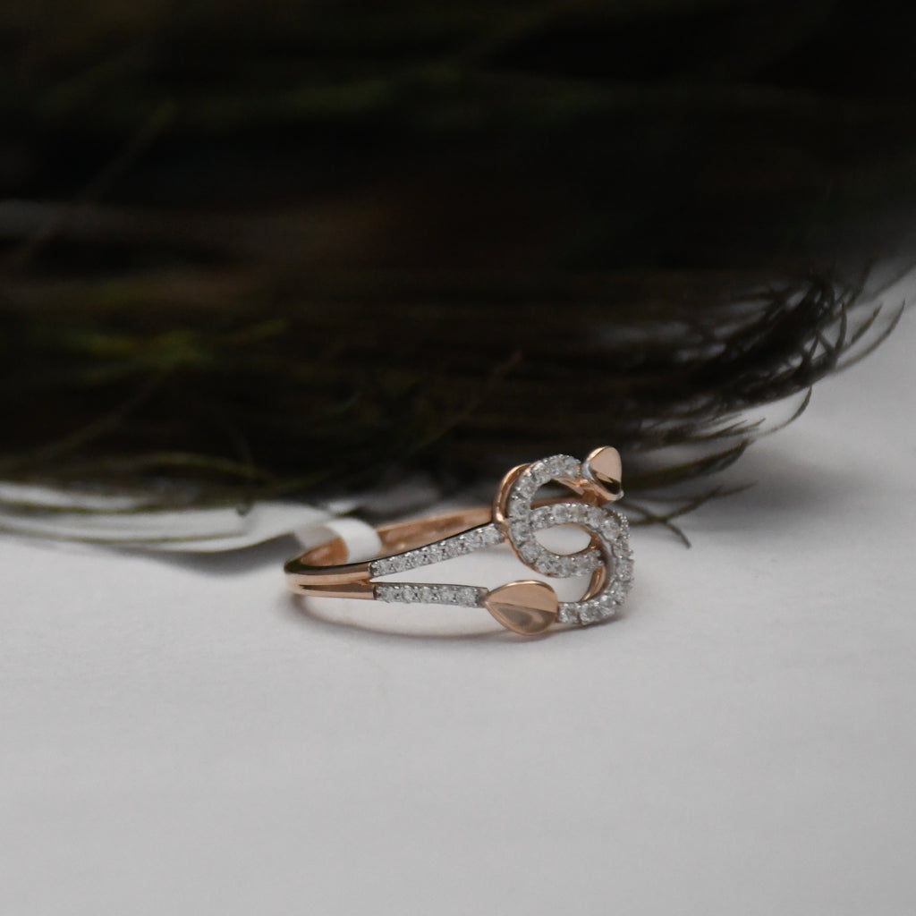 A rose gold ring with pavé-set clear stones and small leaf-shaped accents, photographed on a light surface with dark feathers in the background.